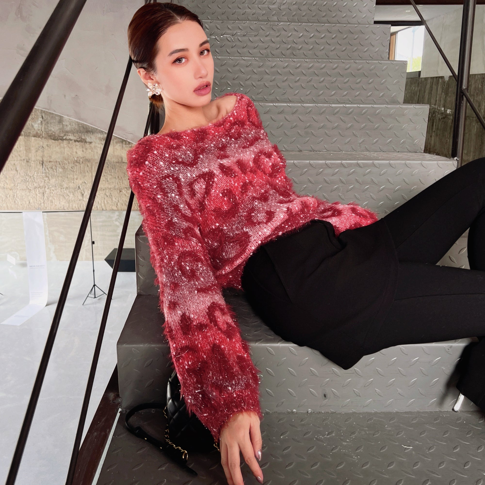 A woman wearing a red, sparkly sweater is sitting on a staircase, looking directly at the camera.