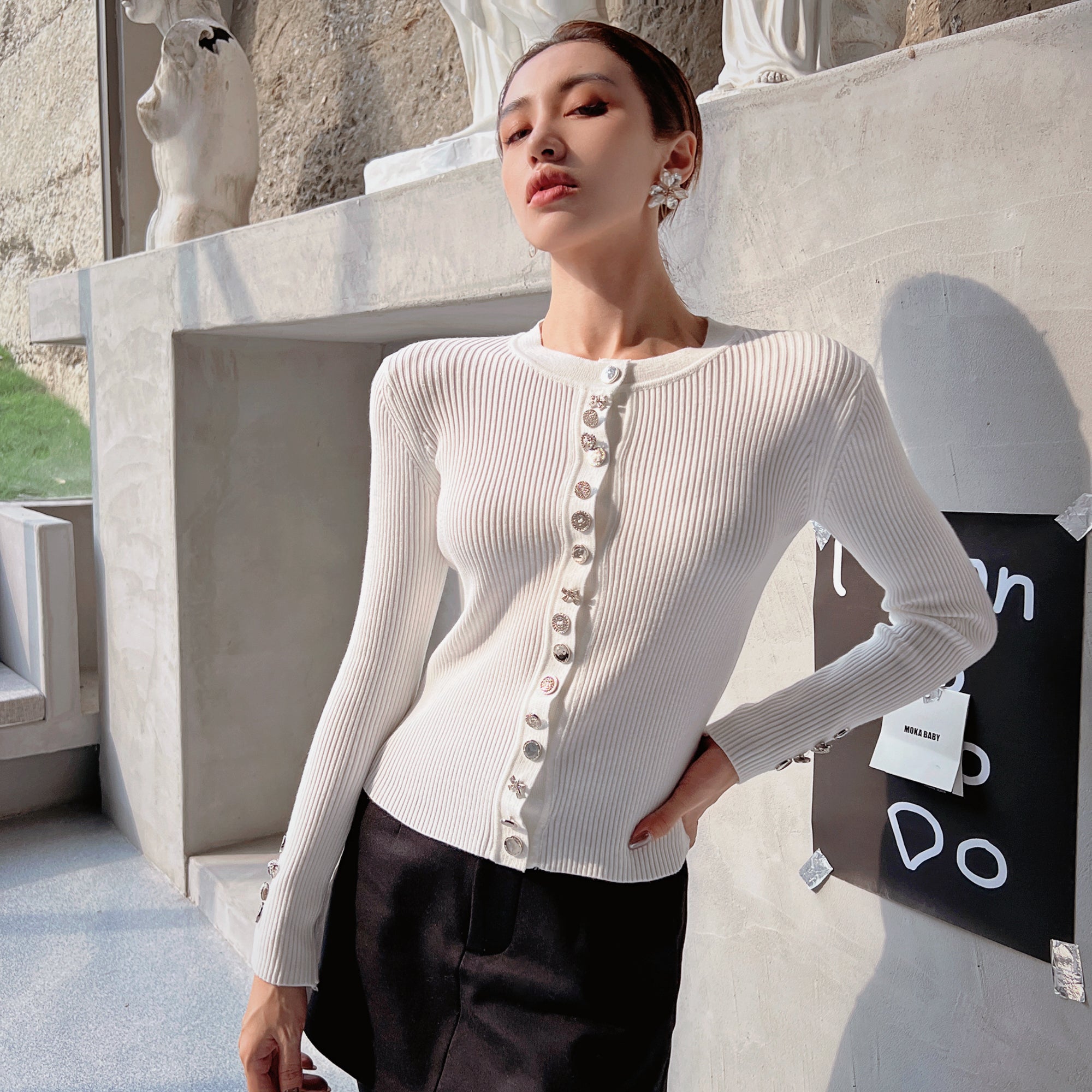A woman wearing a white cardigan with buttons and posing in front of a concrete wall with a sign.