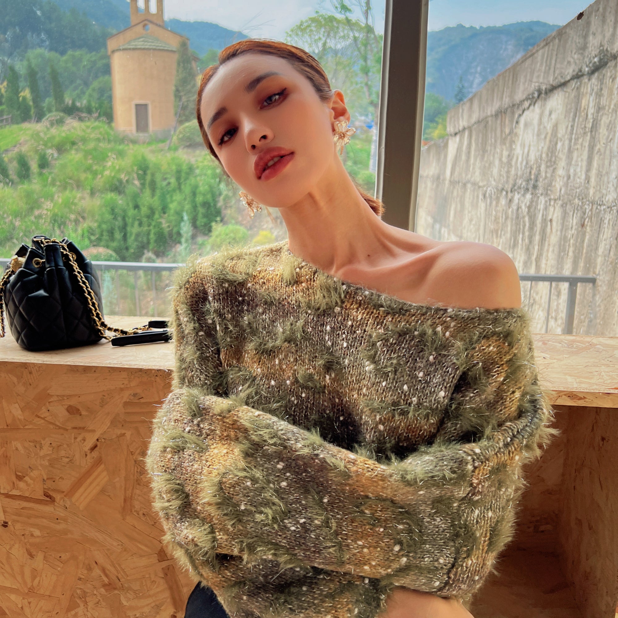 A woman wearing a green and brown patterned sweater stands in front of a window, with a mountainous landscape visible outside.
