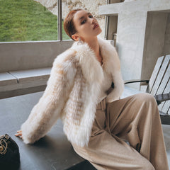 A woman wearing a white fur coat and beige pants sits on a bench, looking off to the side.