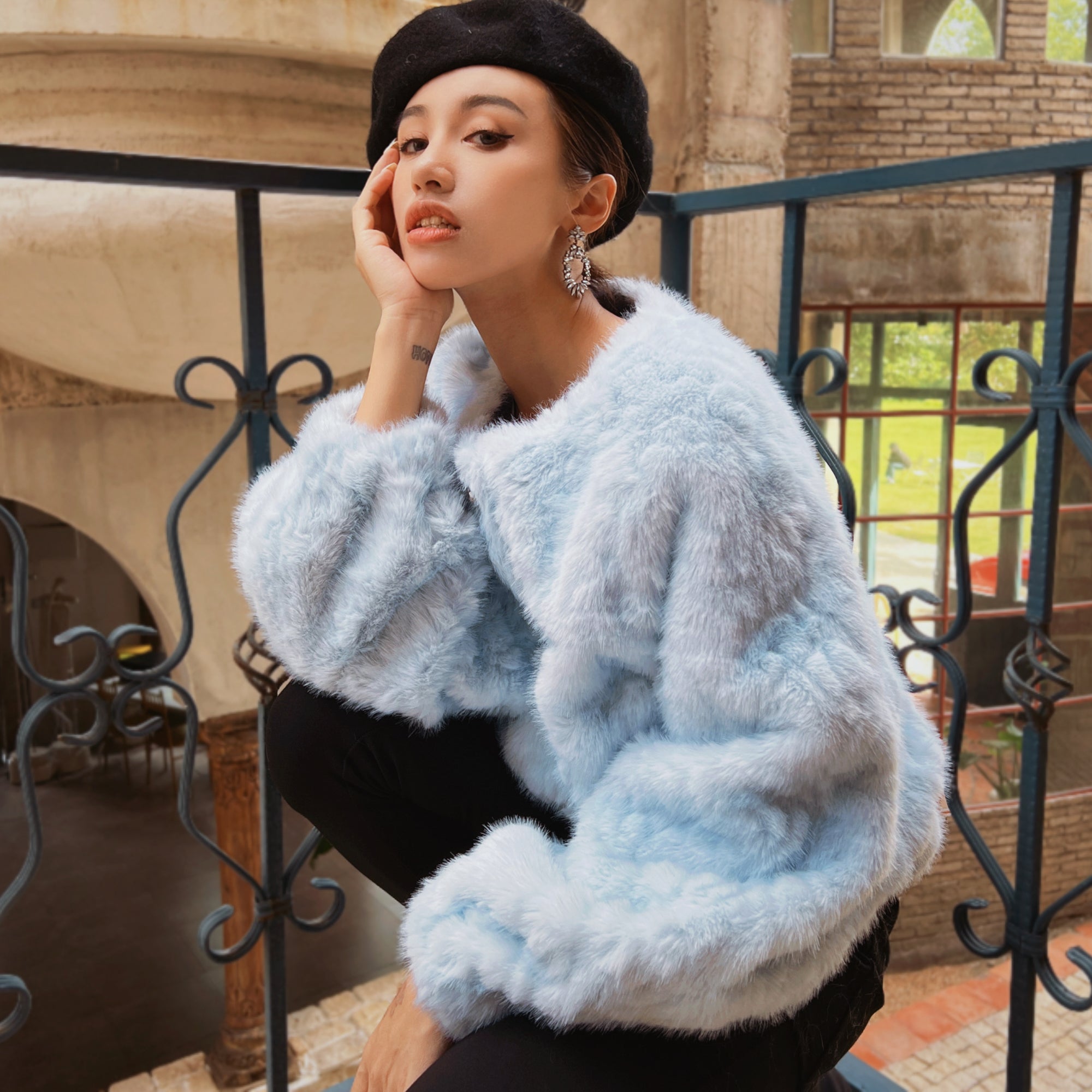 A woman wearing a light blue fur coat and a black hat is posing on a balcony with a wrought iron railing in the background.