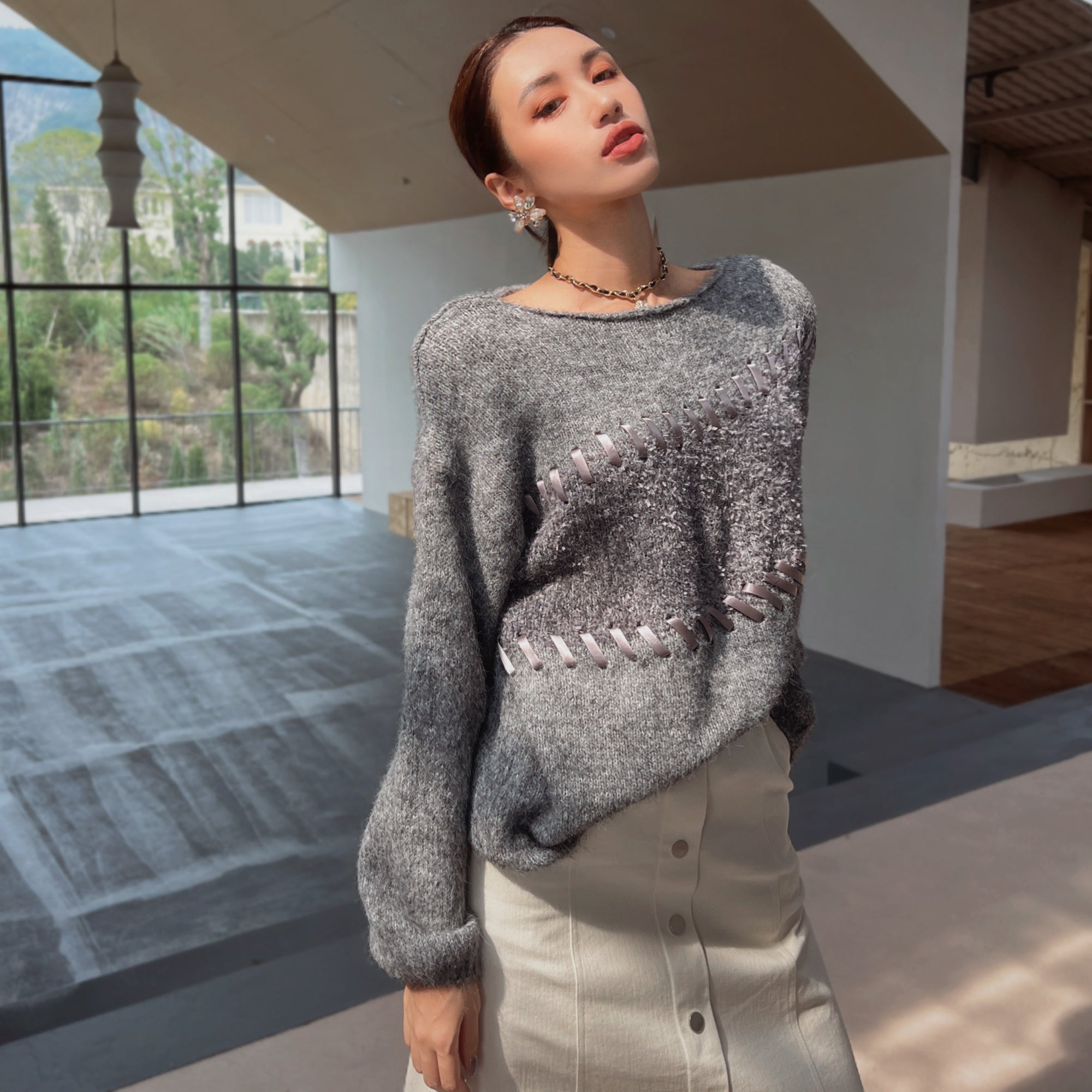 A woman wearing a gray sweater with a lace-like pattern stands in a room with large windows, looking off to the side.