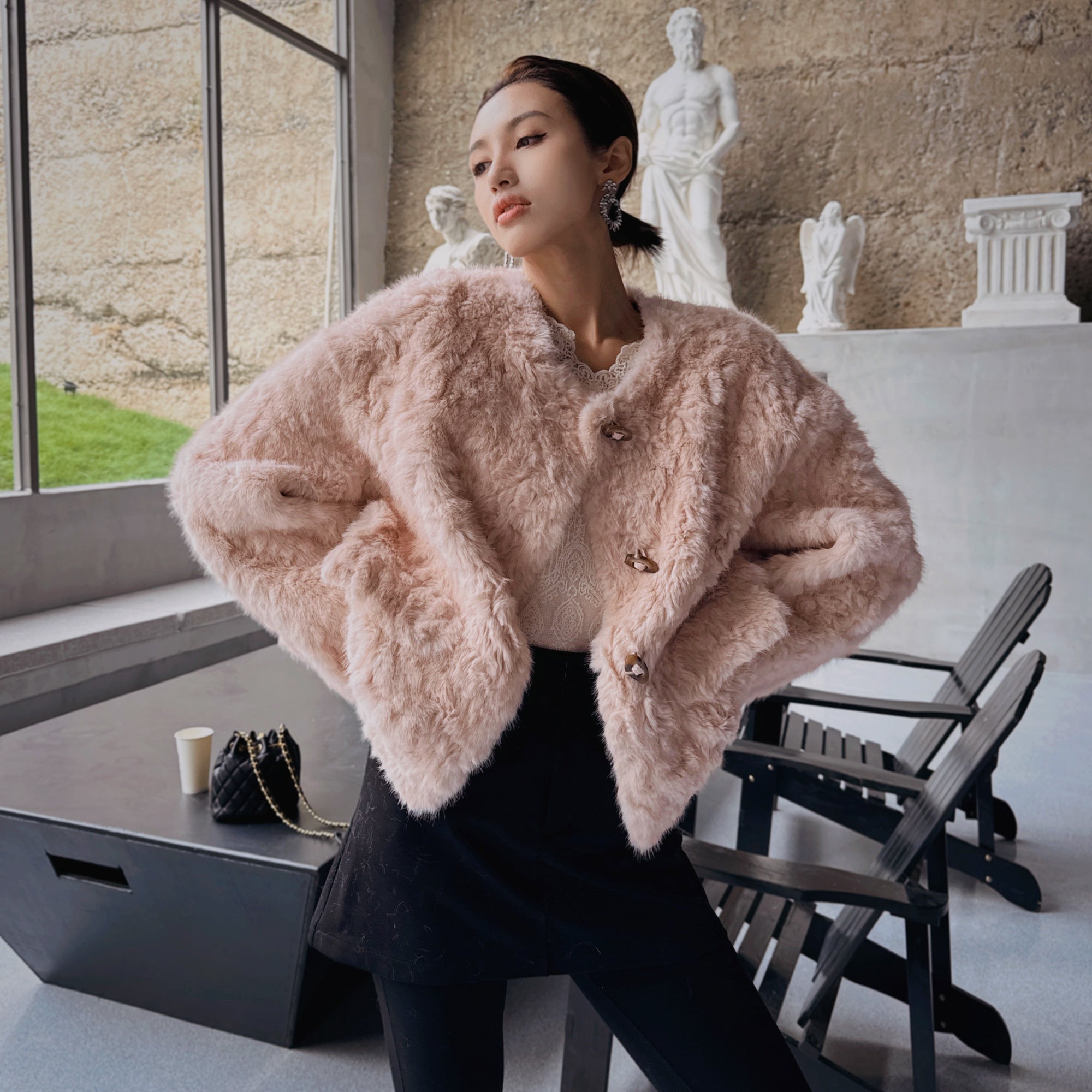 A woman wearing a pink fur coat stands in a room with a window, a statue, and a black chair.