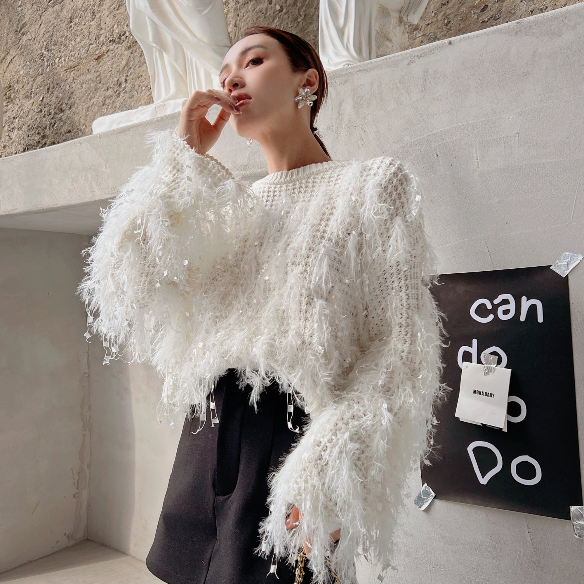 A woman wearing a white sweater with fringe and a black skirt is posing in front of a white wall with a black sign that says "can do do".