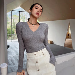 A woman wearing a gray sweater and white pants stands in a room with a large window, looking off to the side.