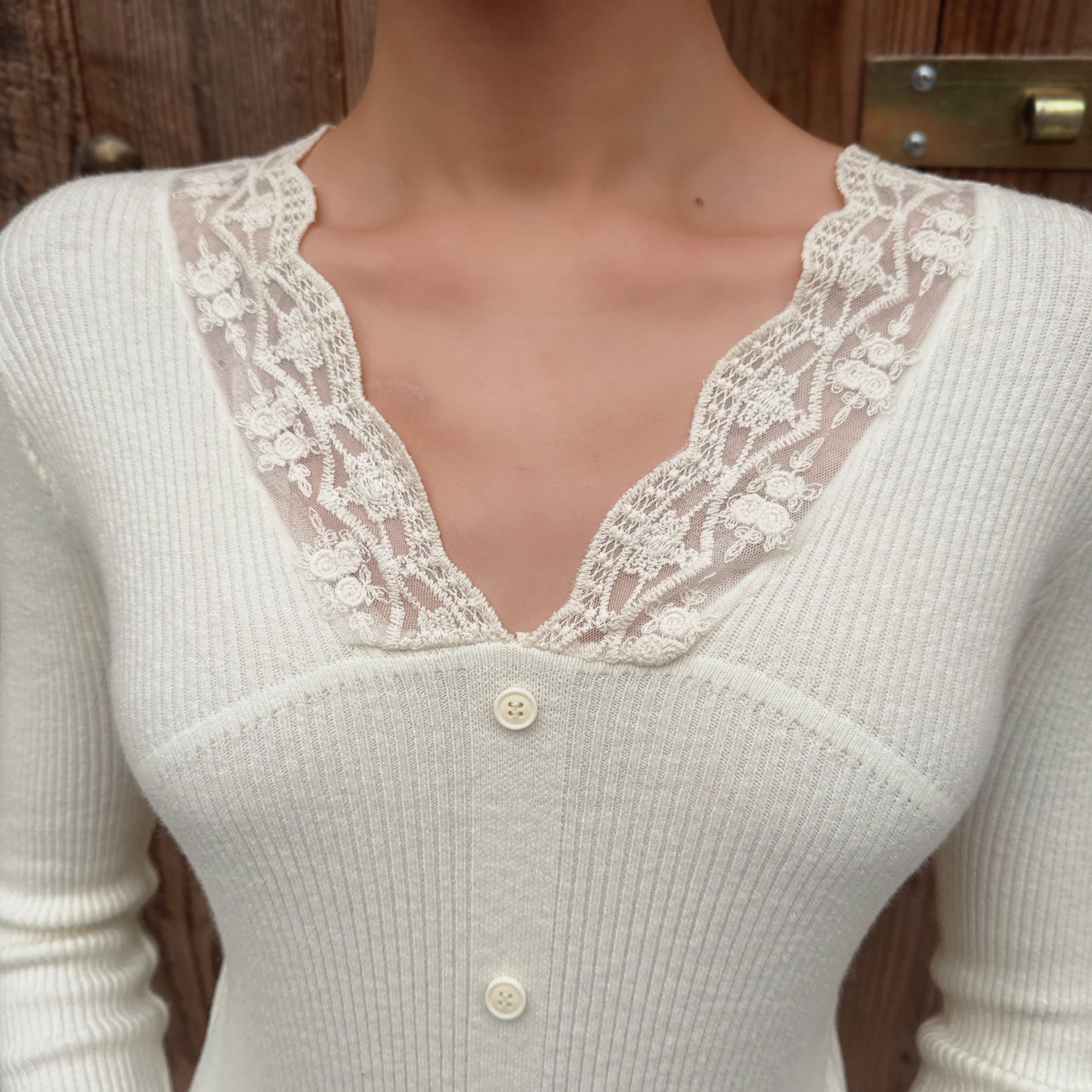 A close-up of a woman wearing a white lace-trimmed cardigan with buttons, standing in front of a wooden door.