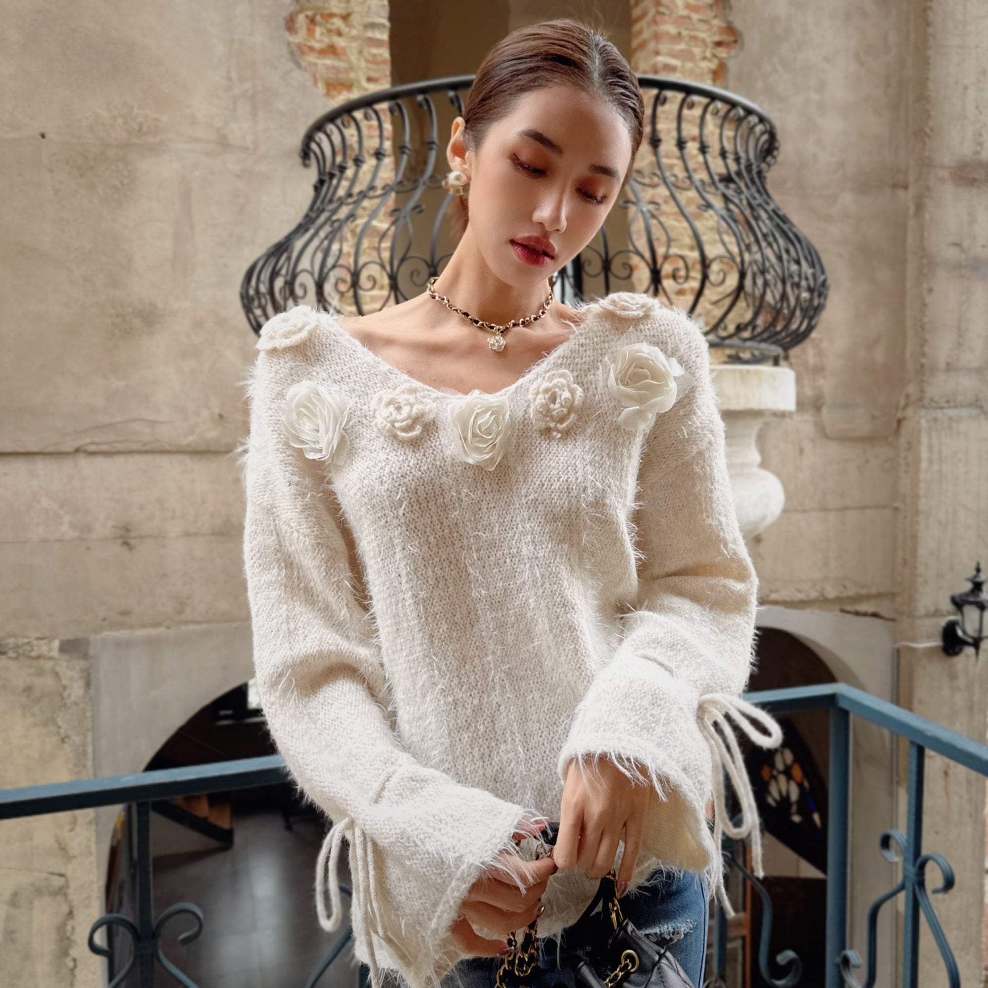 A woman wearing a white sweater with flower embellishments stands in front of a wrought iron balcony, holding a purse.