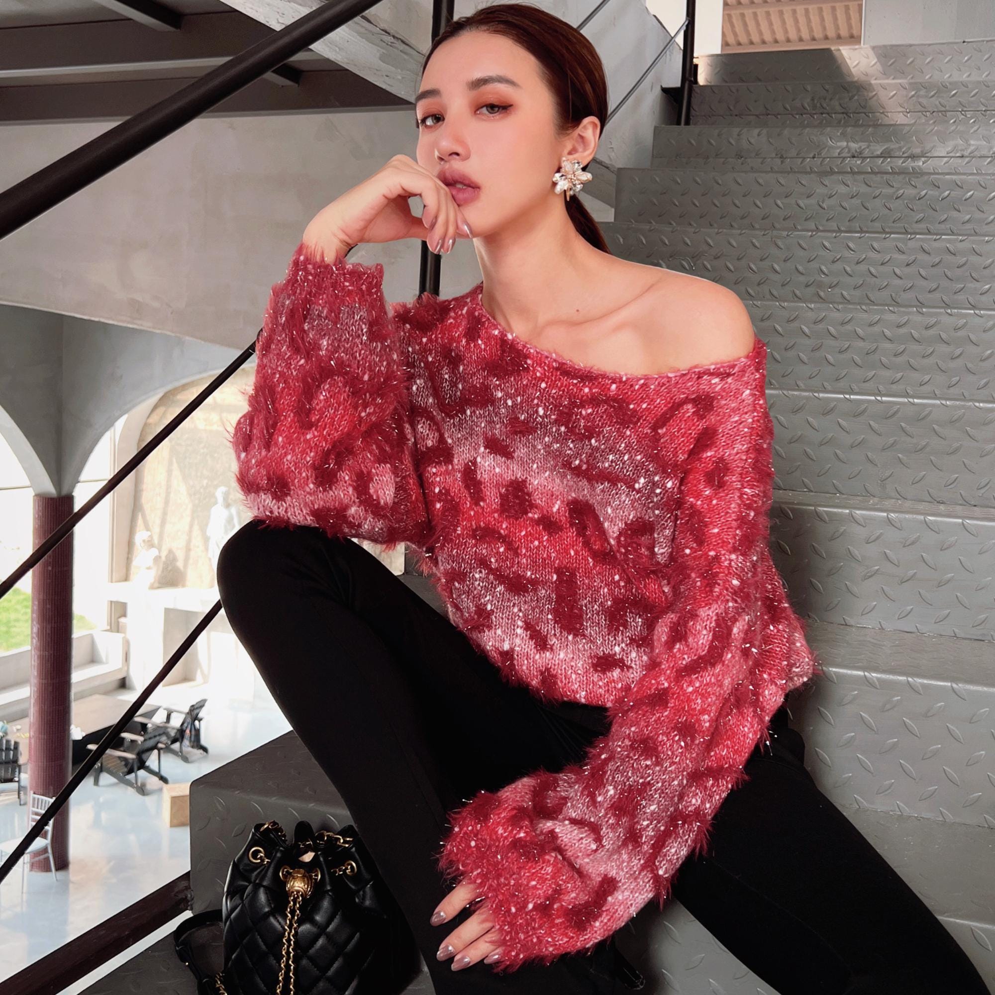 A woman wearing a red and pink patterned sweater and black pants is sitting on a staircase, with a handbag next to her.