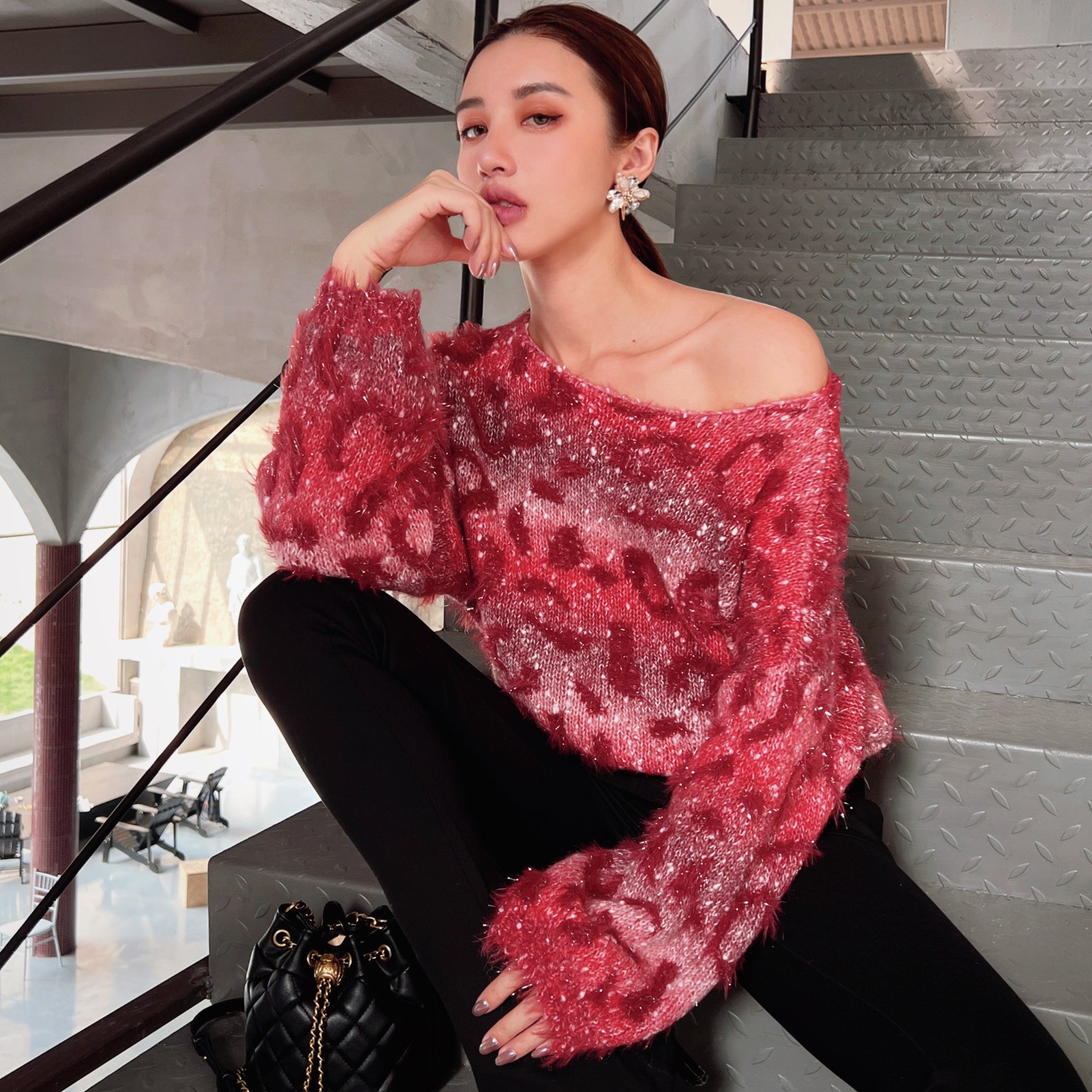 A woman wearing a red and pink patterned sweater and black pants is sitting on a staircase, with a handbag next to her.