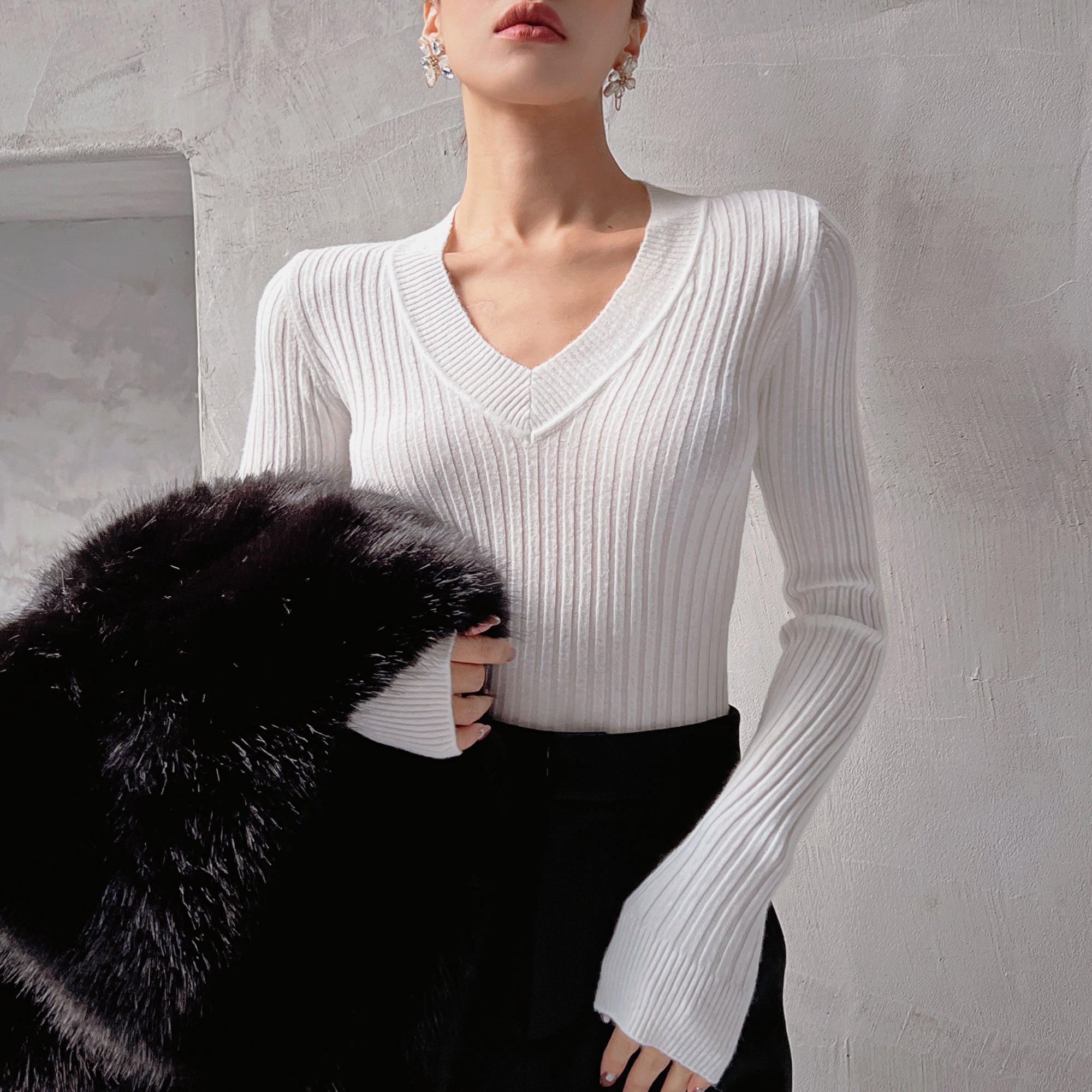A woman wearing a white sweater and black pants stands against a white wall, holding a black fur coat.