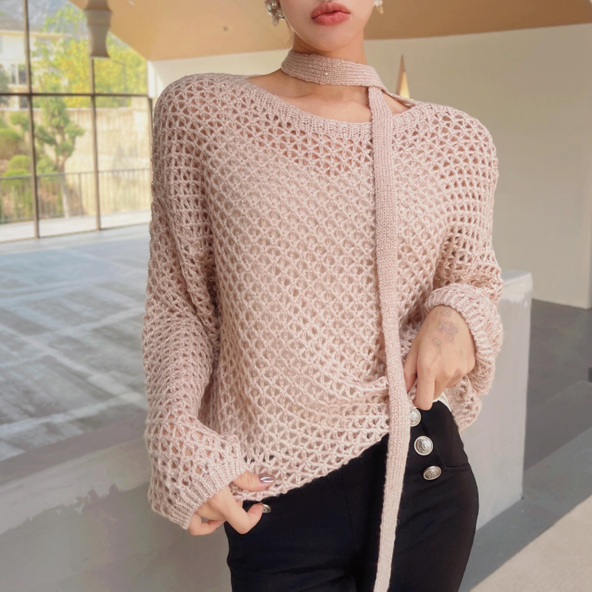 A woman wearing a pink knitted sweater with a choker necktie, posing in an indoor setting with a window in the background.