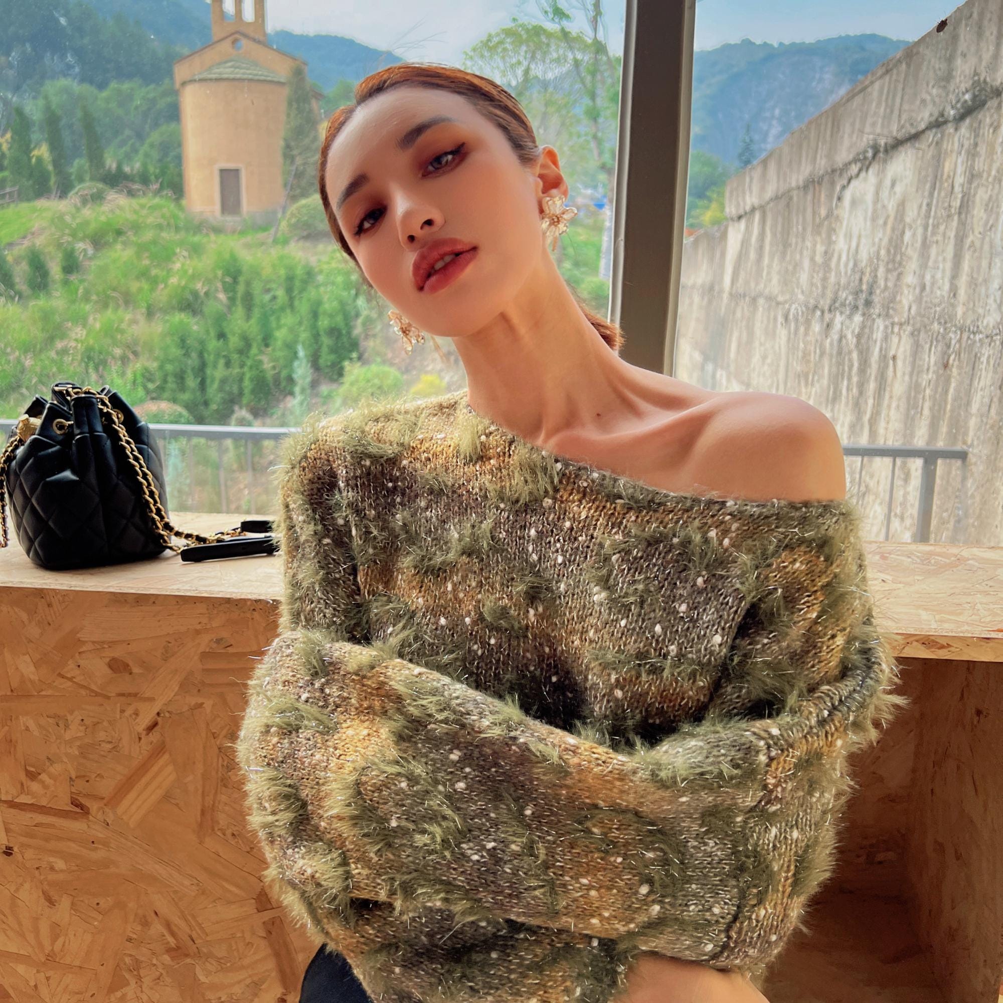 A woman wearing a green and brown patterned sweater stands in front of a window, with a mountainous landscape visible outside.