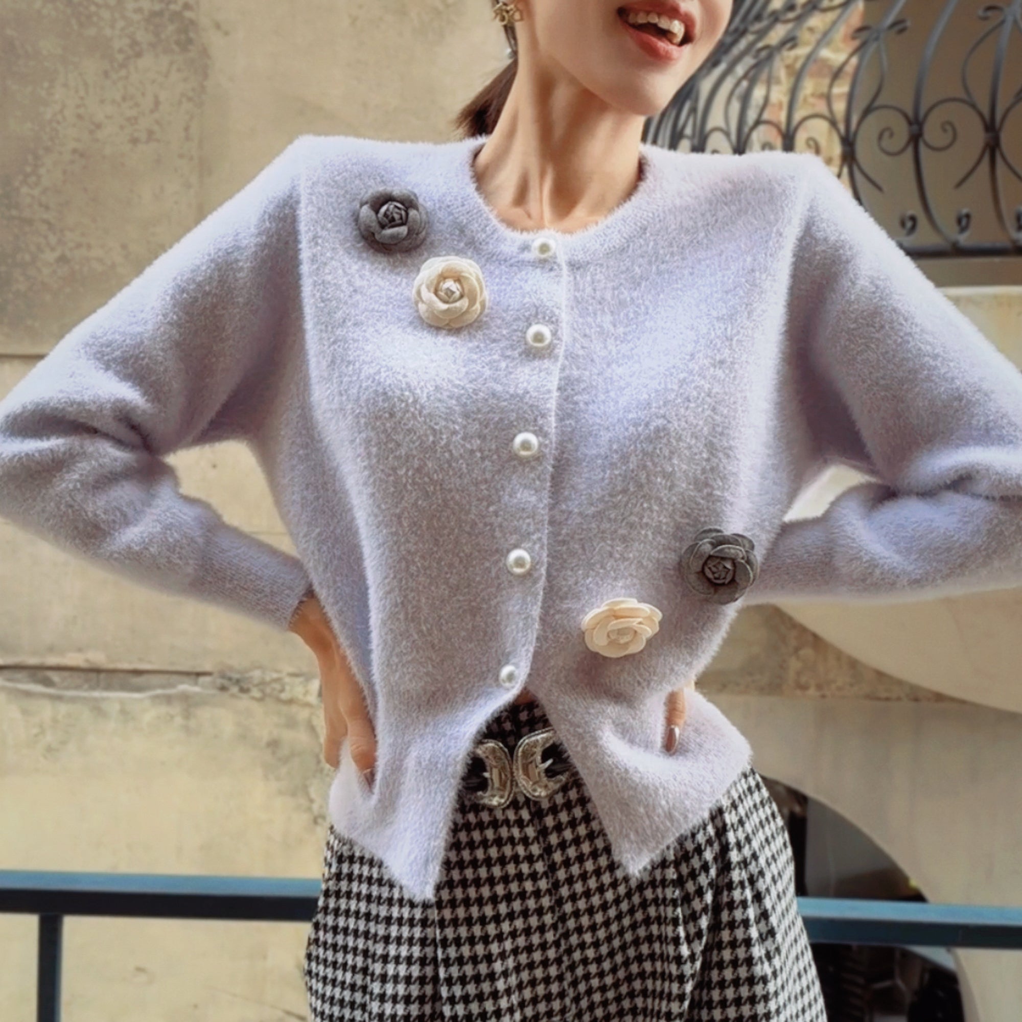 A woman wearing a white cardigan with floral embellishments on the front, posing with her hands on her hips.