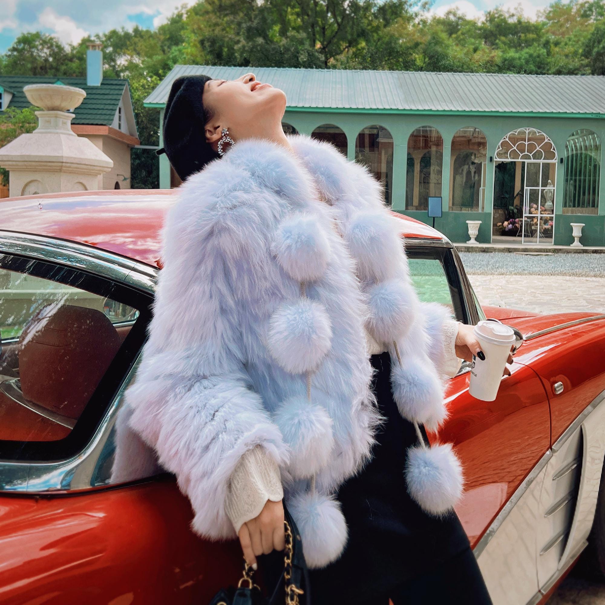 A woman in a white fur coat stands next to a red car, holding a coffee cup and looking up at the sky.