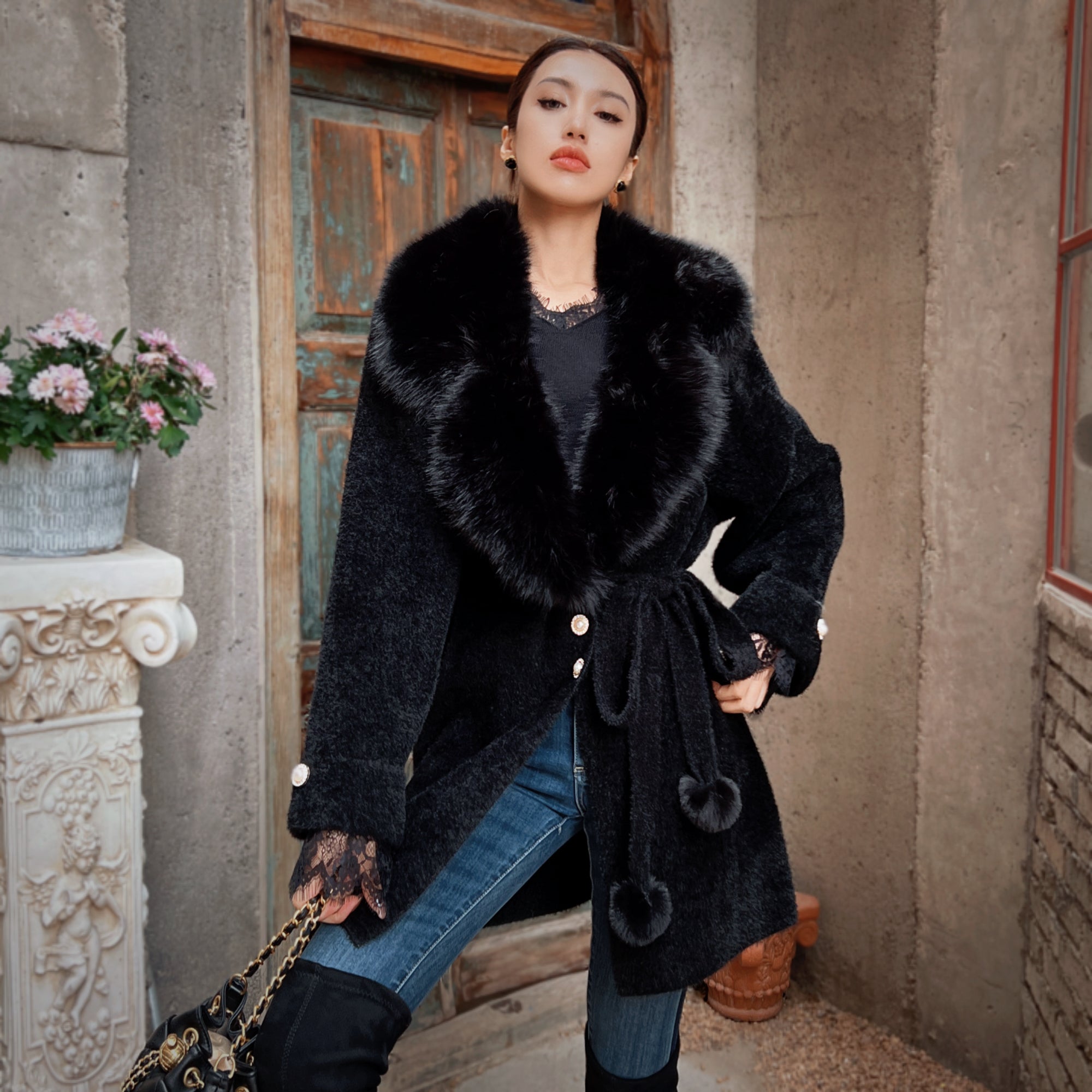 A woman wearing a black fur coat and holding a purse stands in front of a doorway with a floral planter.