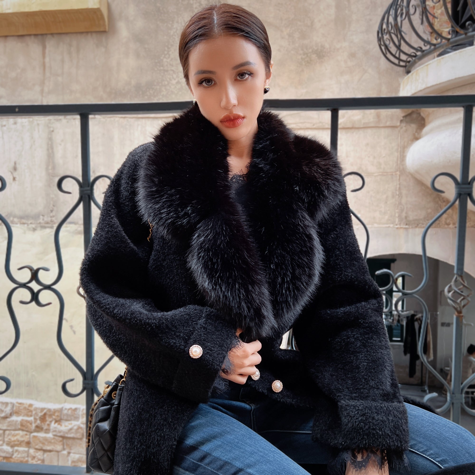 A woman wearing a black fur coat with a fur collar sits on a bench, looking directly at the camera with a serious expression.