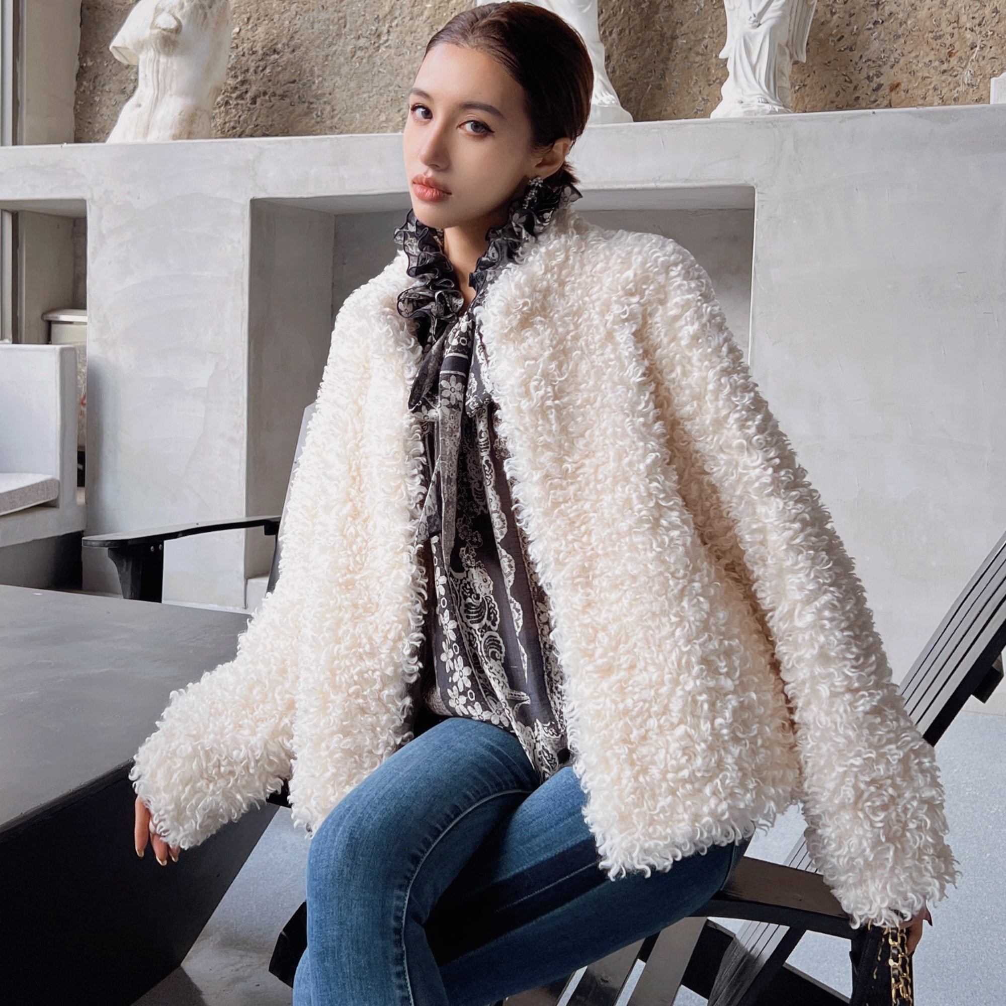 A woman wearing a white fluffy coat and a patterned scarf is sitting on a chair in an indoor setting.