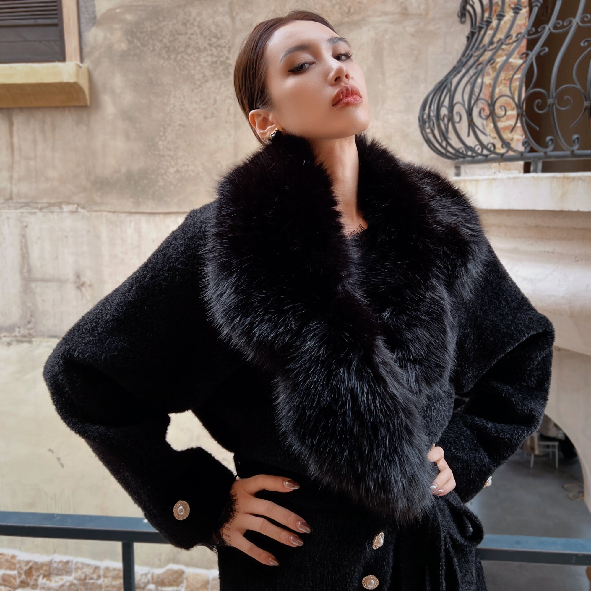 A woman wearing a black fur coat stands confidently with her hands on her hips, posing against a stone wall.