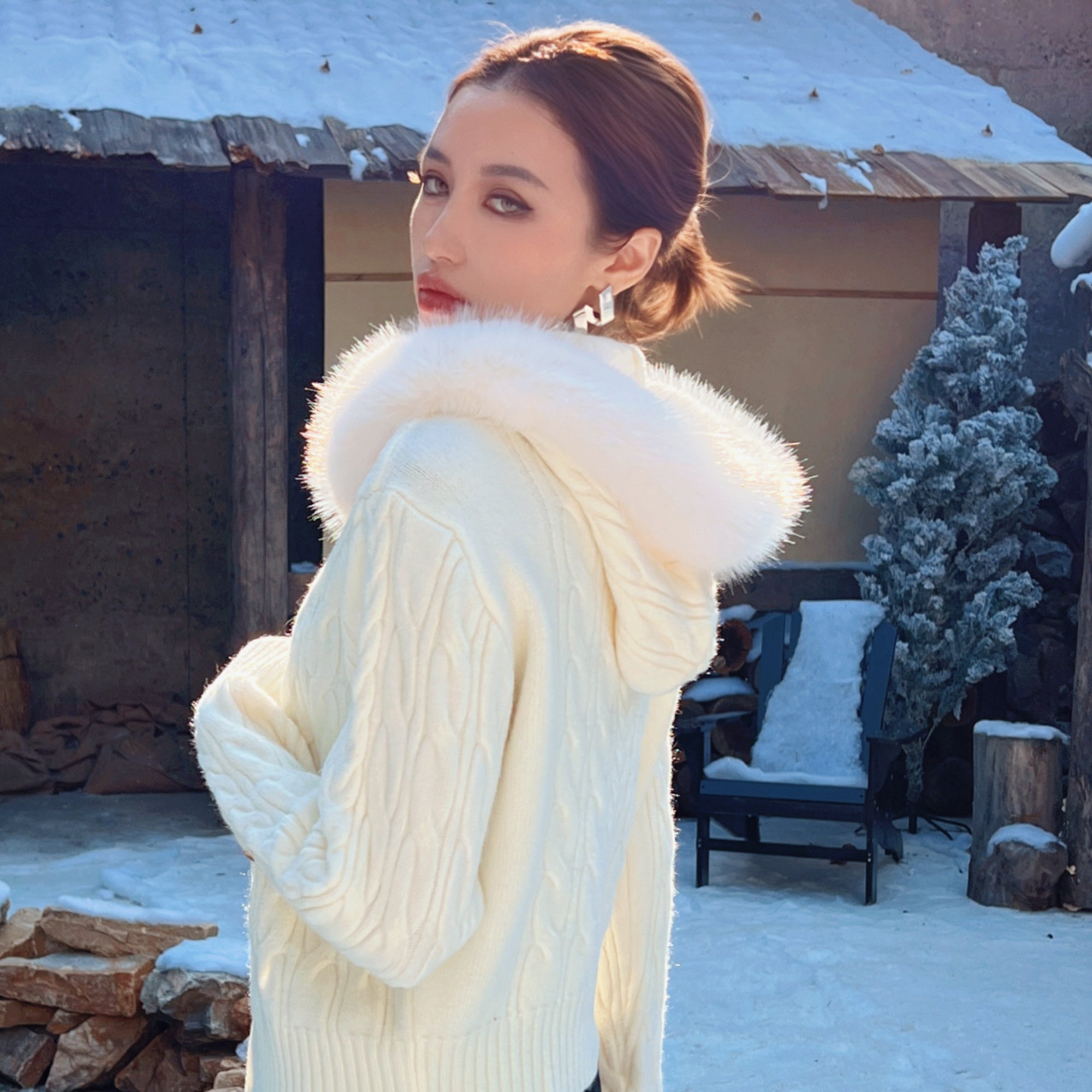 A woman in a white sweater with a fur collar stands in front of a snowy wooden structure, with a chair and a tree visible in the background.