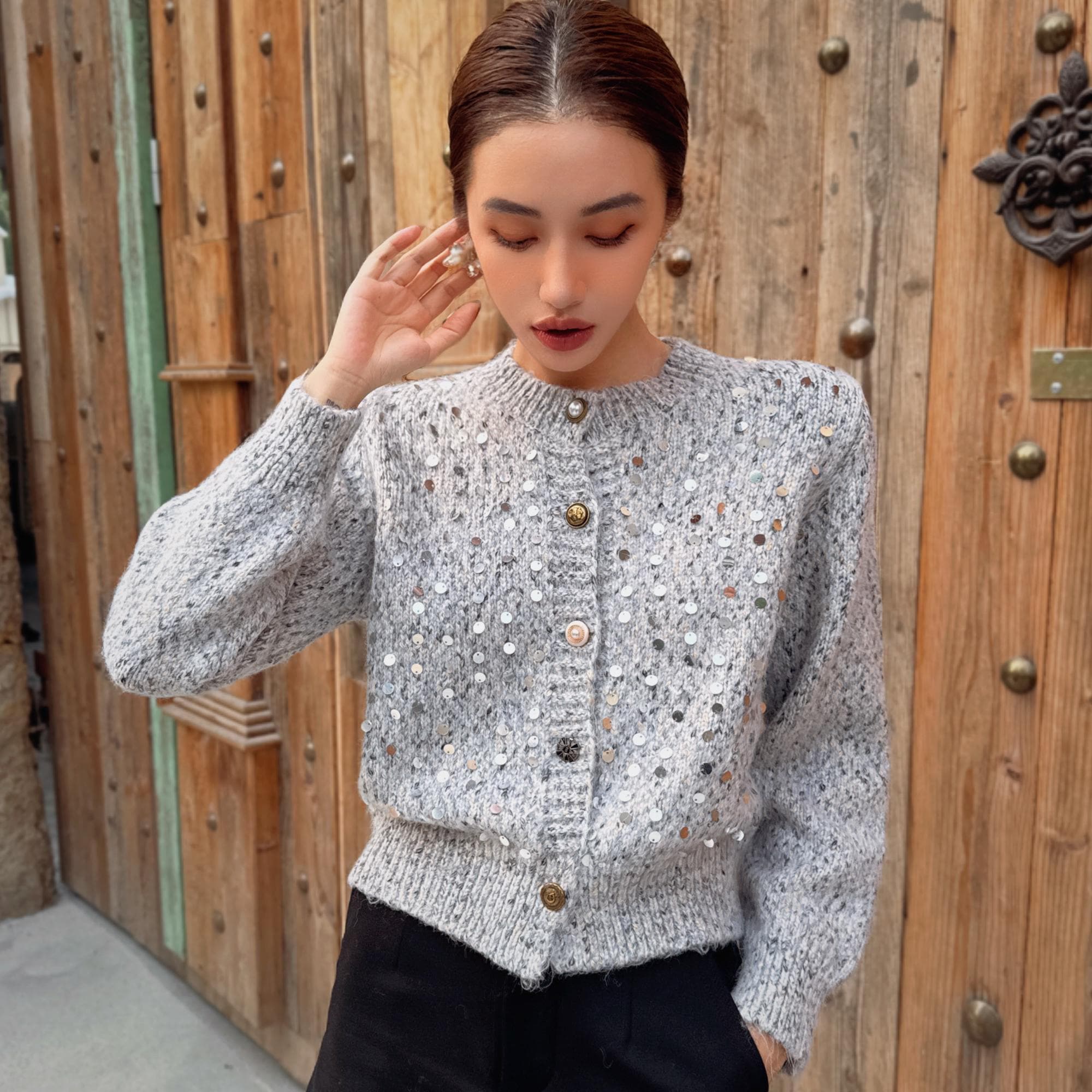 A woman wearing a gray sweater with silver buttons stands in front of a wooden door.