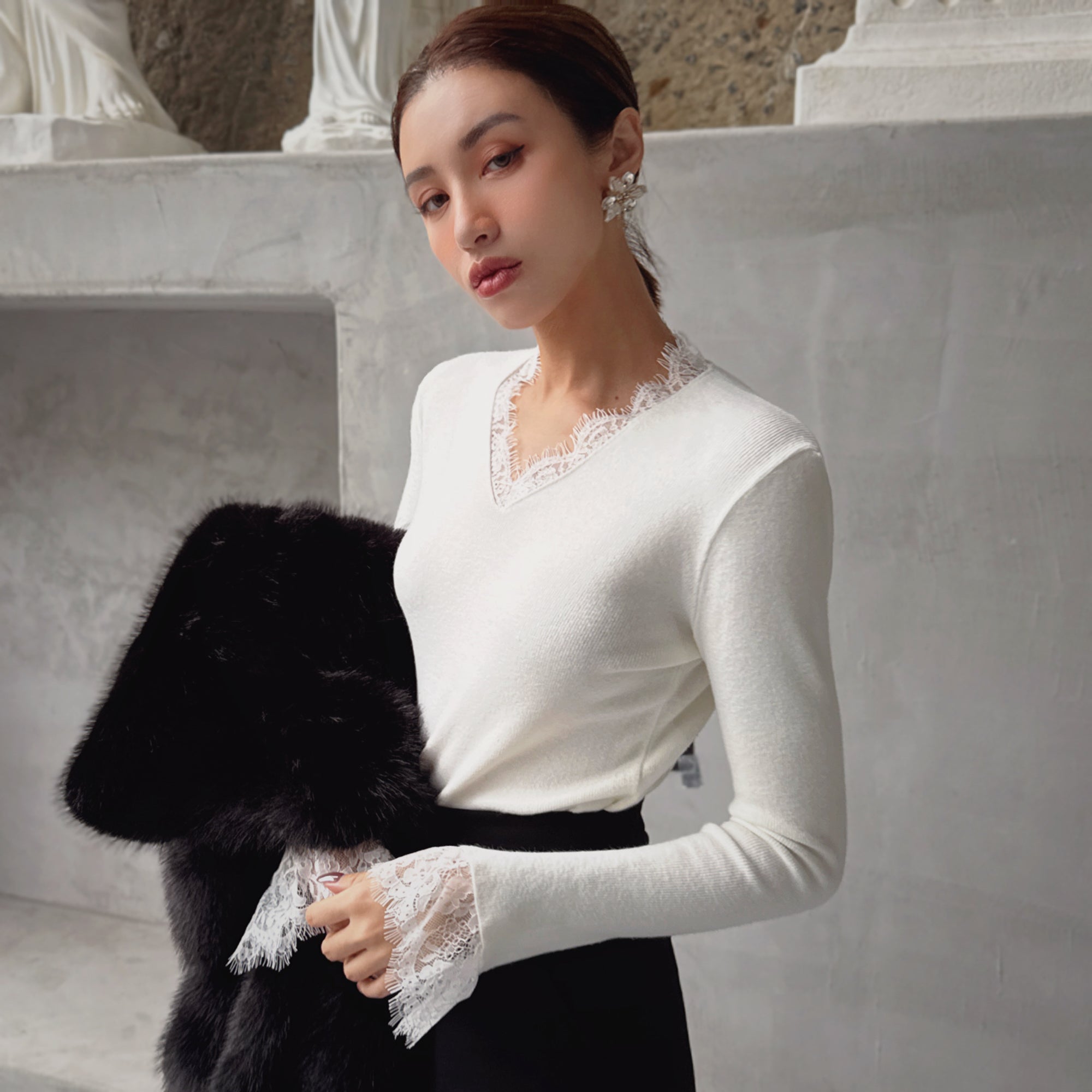 A woman wearing a white sweater with lace trim and holding a black fur coat stands in front of a stone wall.