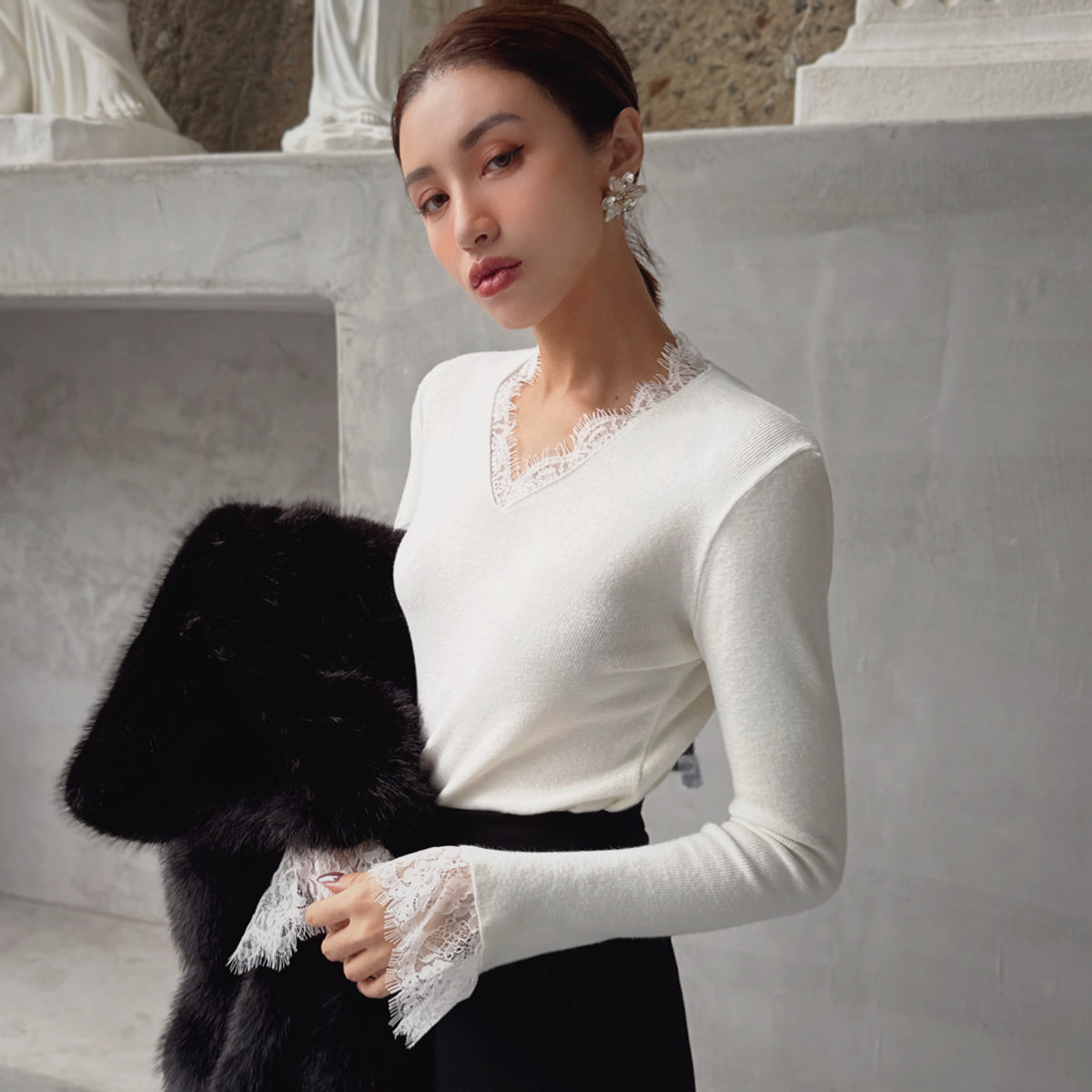 A woman wearing a white sweater with lace trim and holding a black fur coat stands in front of a stone wall.