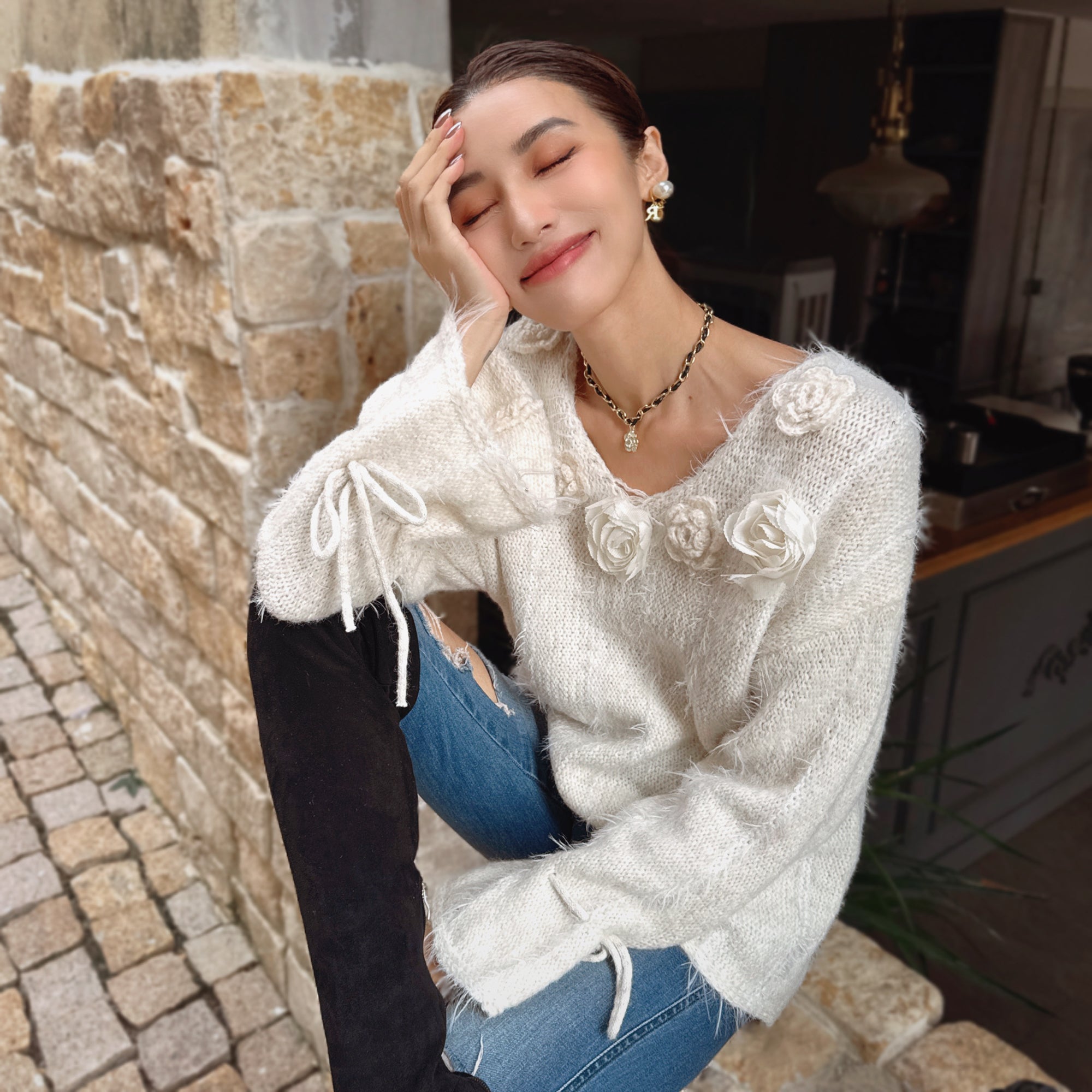 A woman wearing a white sweater with flower designs and a necklace is sitting on a stone wall, with her eyes closed and her hand resting on her head.