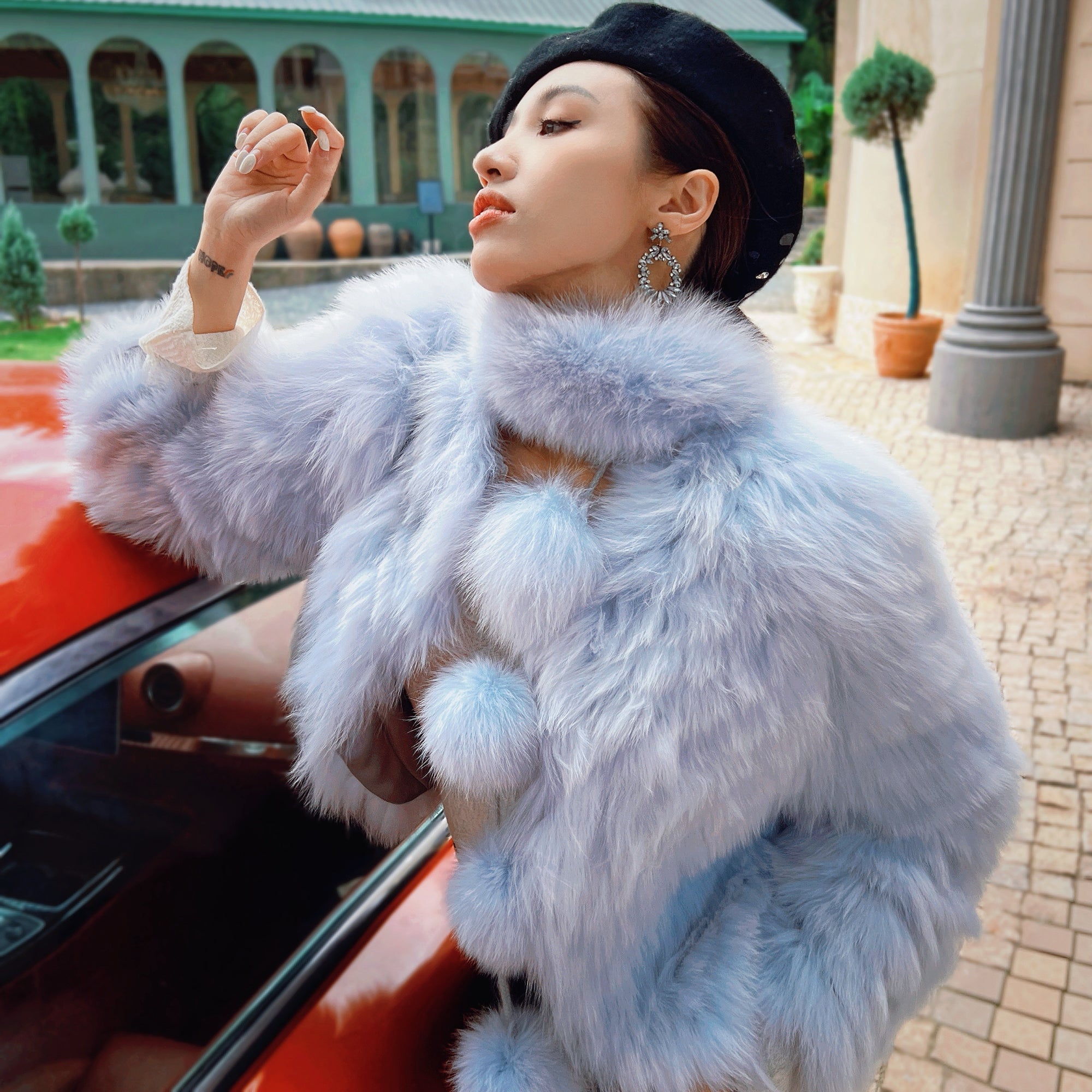 A woman in a blue fur coat and black hat stands next to a red car, with a building and potted plants in the background.