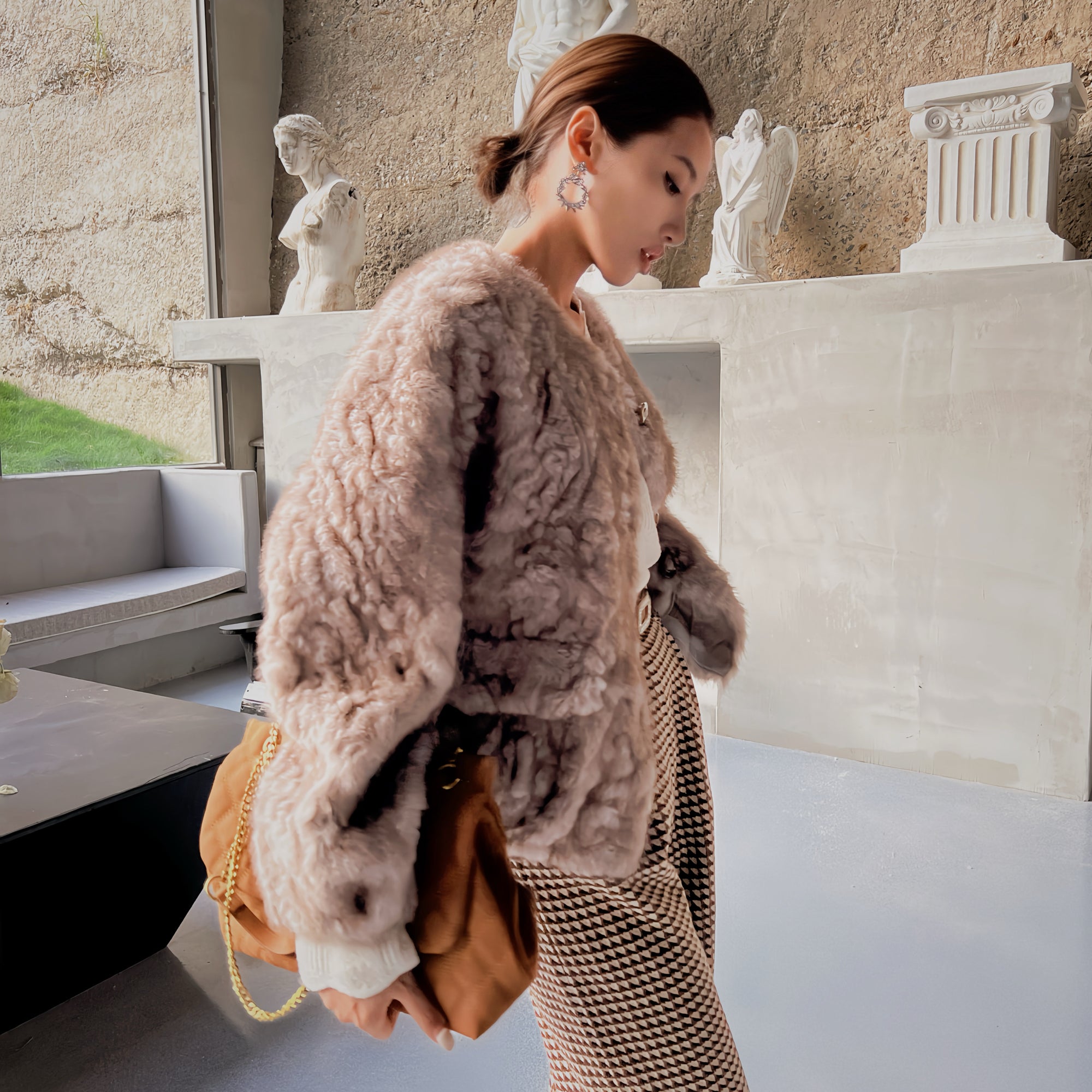 A woman wearing a fur coat and carrying a brown purse is standing in a room with a stone wall and statues in the background.