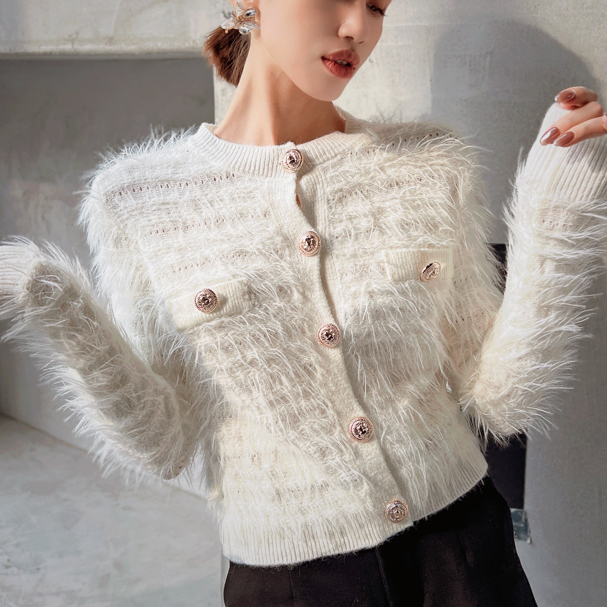A woman wearing a white, furry cardigan with buttons, posing in front of a white wall.