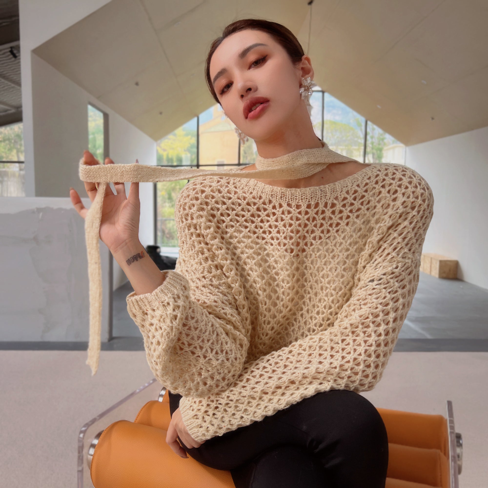 A woman wearing a beige sweater and holding a scarf around her neck is sitting on an orange chair in a room with large windows.