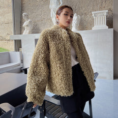 A woman wearing a beige furry coat stands in a room with a stone wall and statues in the background.