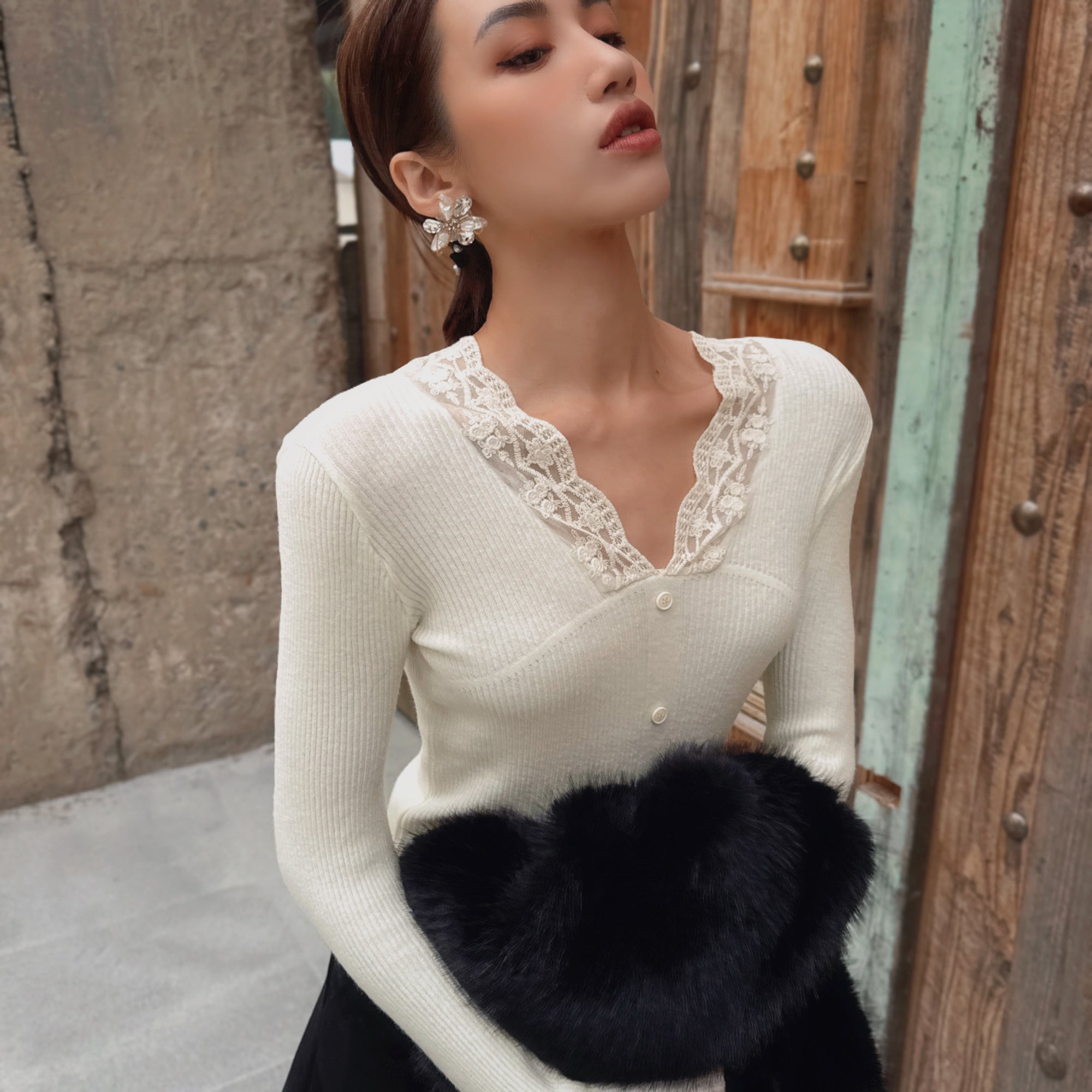 A woman wearing a white lace top and holding a black fur coat stands in front of a wooden wall.