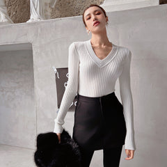 A woman wearing a white sweater and black skirt stands in front of a white wall, holding a black fur coat.