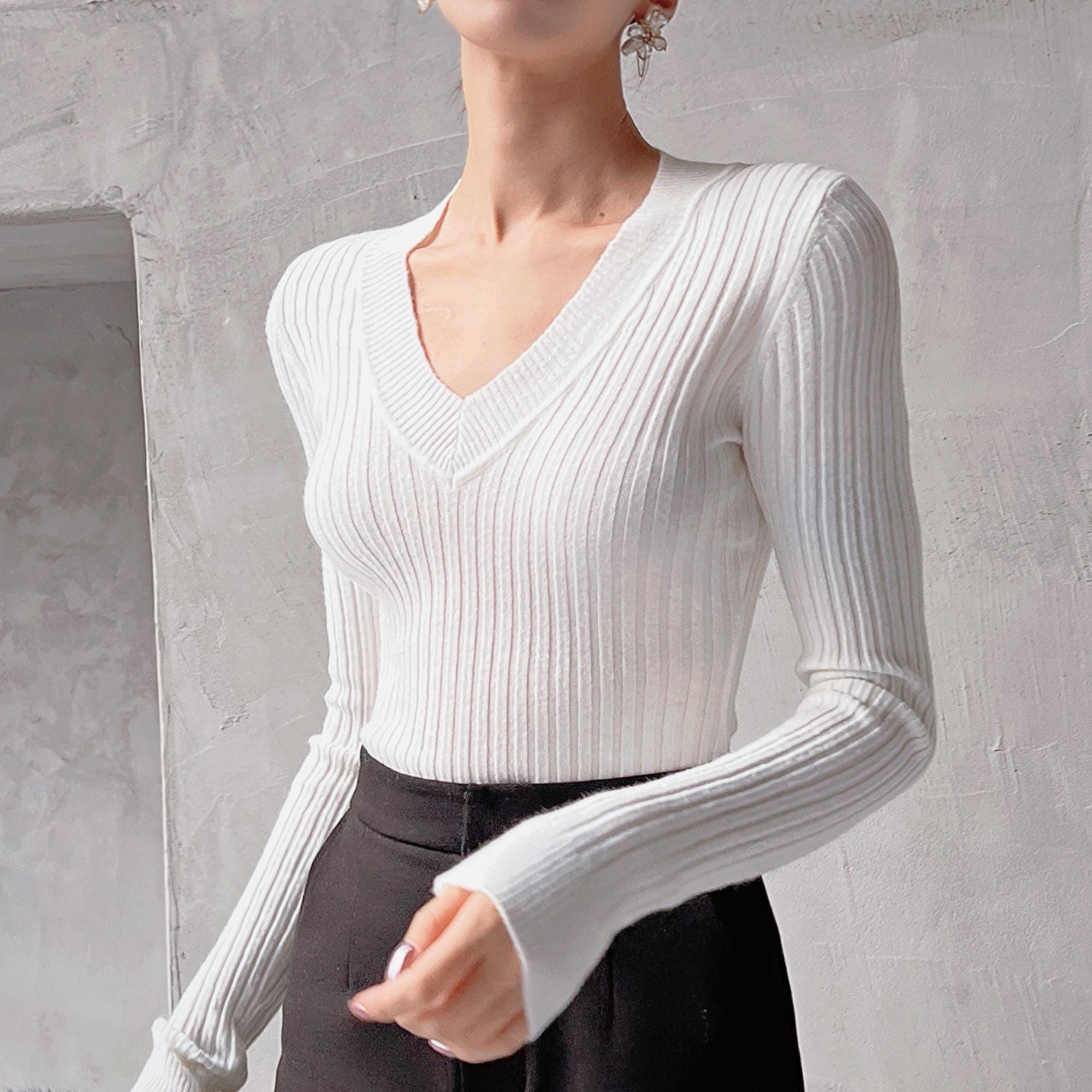 A woman wearing a white v-neck sweater with long sleeves, standing against a plain white wall.