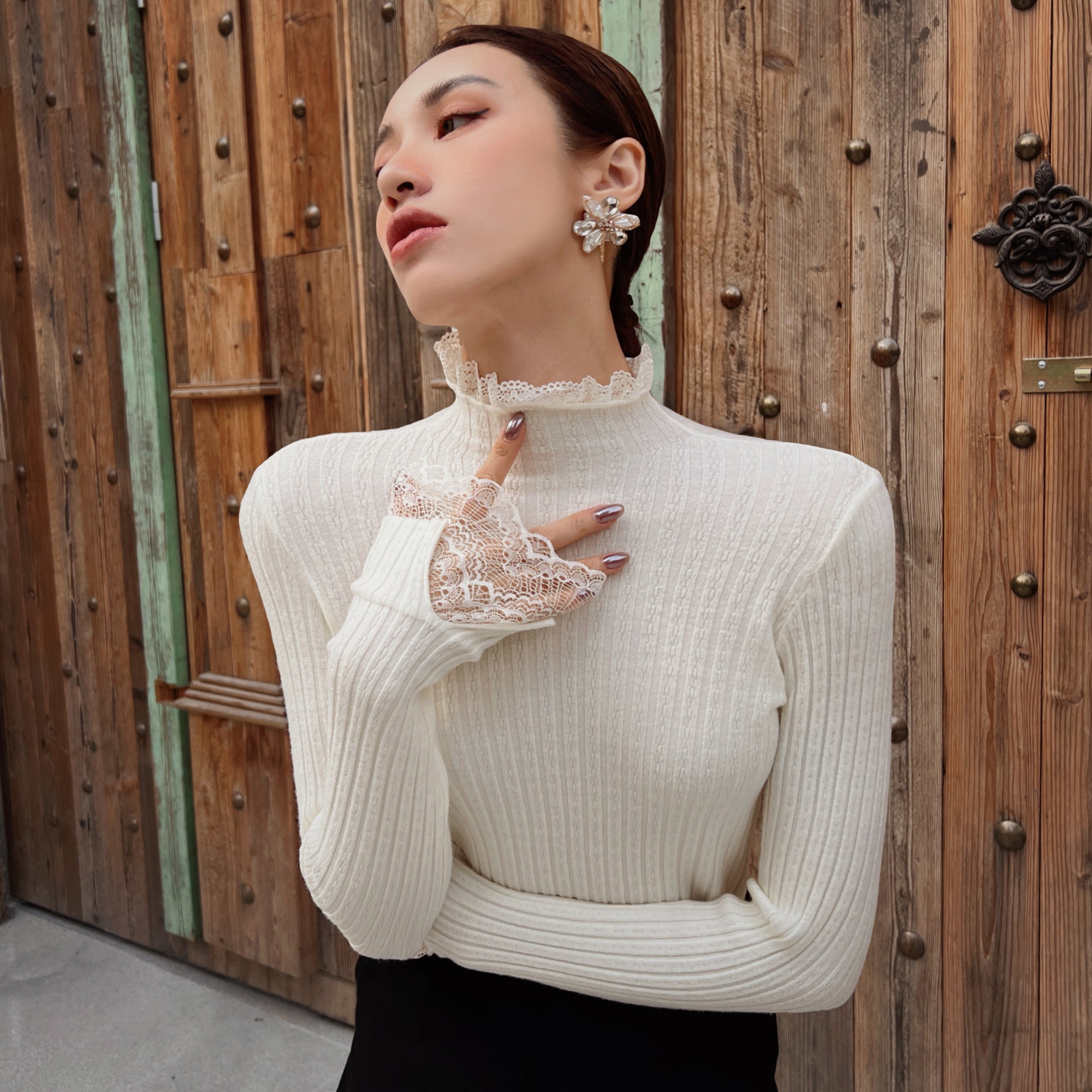 A woman wearing a white lace turtleneck sweater and a pair of large earrings stands in front of a wooden door.