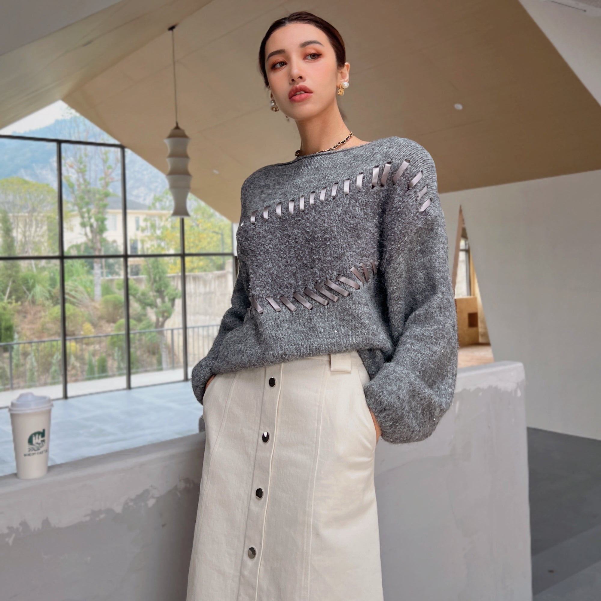 A woman wearing a gray sweater with a unique design and a white skirt stands in a room with a large window overlooking a mountainous landscape.