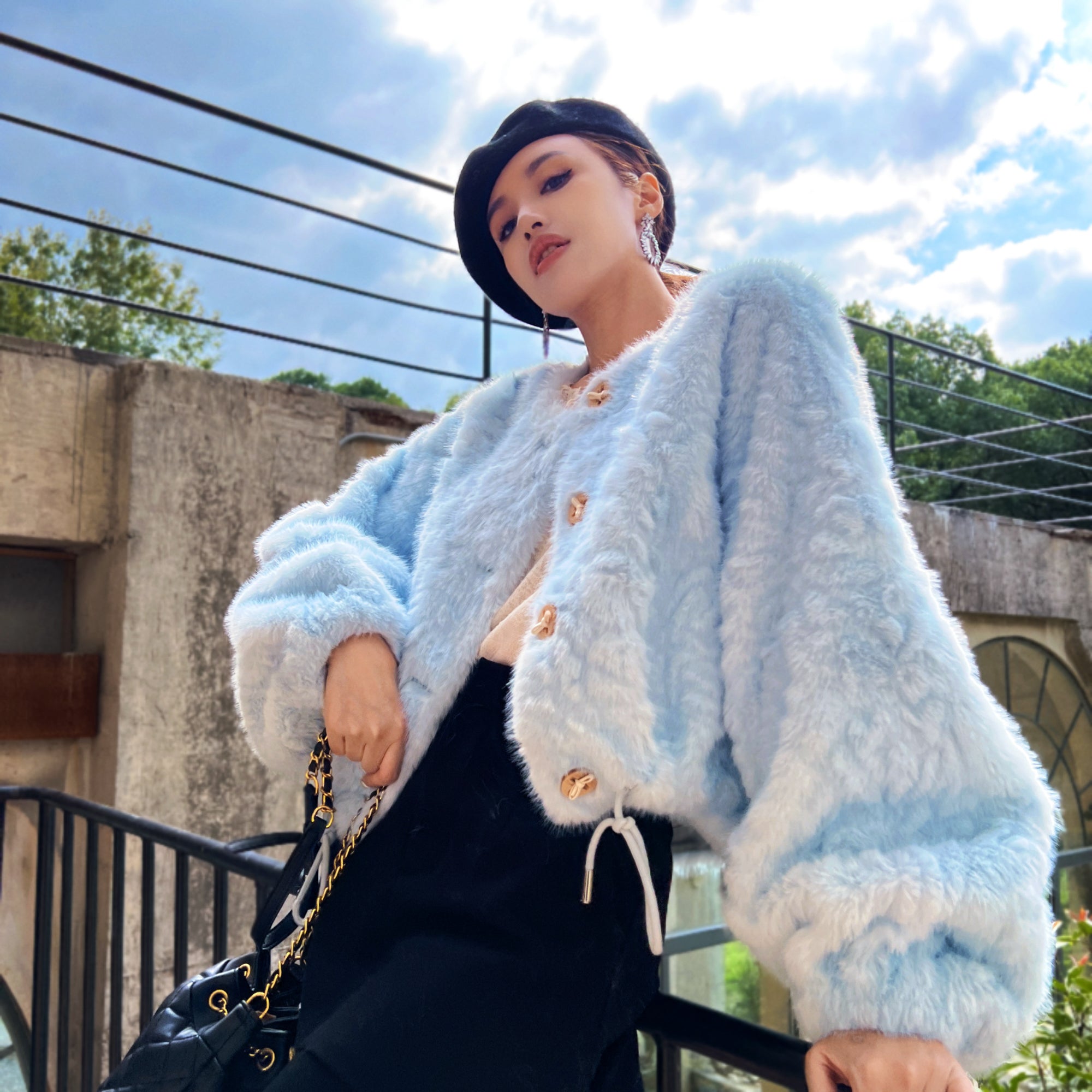 A woman wearing a light blue oversized coat and a black skirt is posing outdoors against a backdrop of a building and trees.