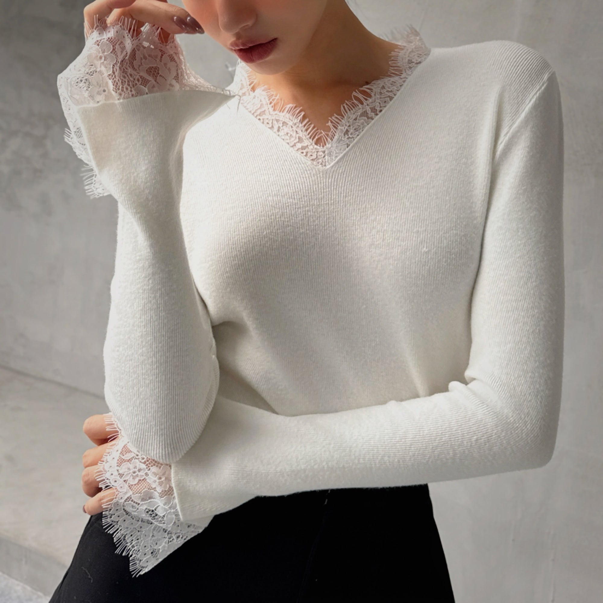 A woman wearing a white sweater with lace trim and a black skirt, posing with her hand on her chin.