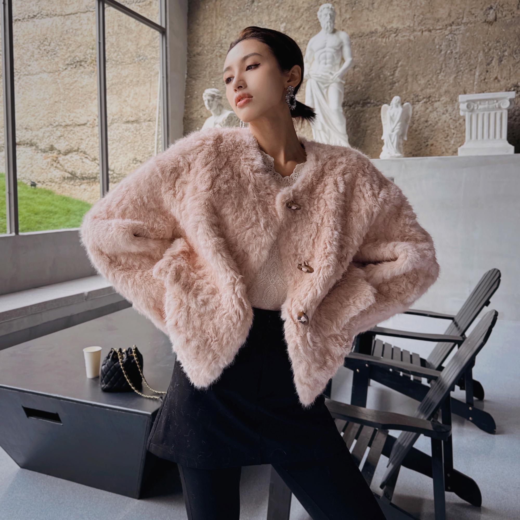 A woman wearing a pink fur coat stands in a room with a window, a statue, and a black chair.