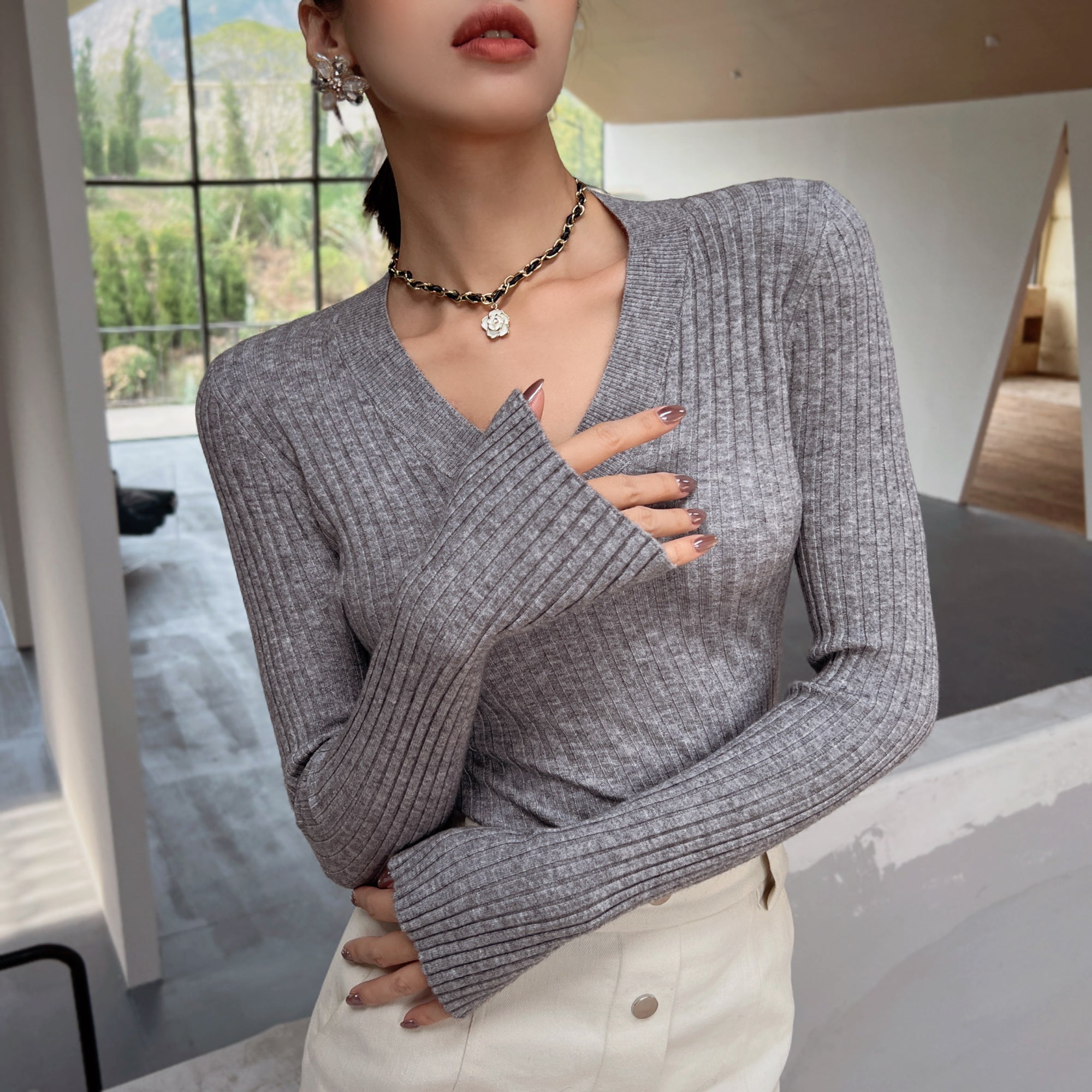 A woman wearing a gray sweater and white pants stands in a room with a window, her hands tucked into her pockets.