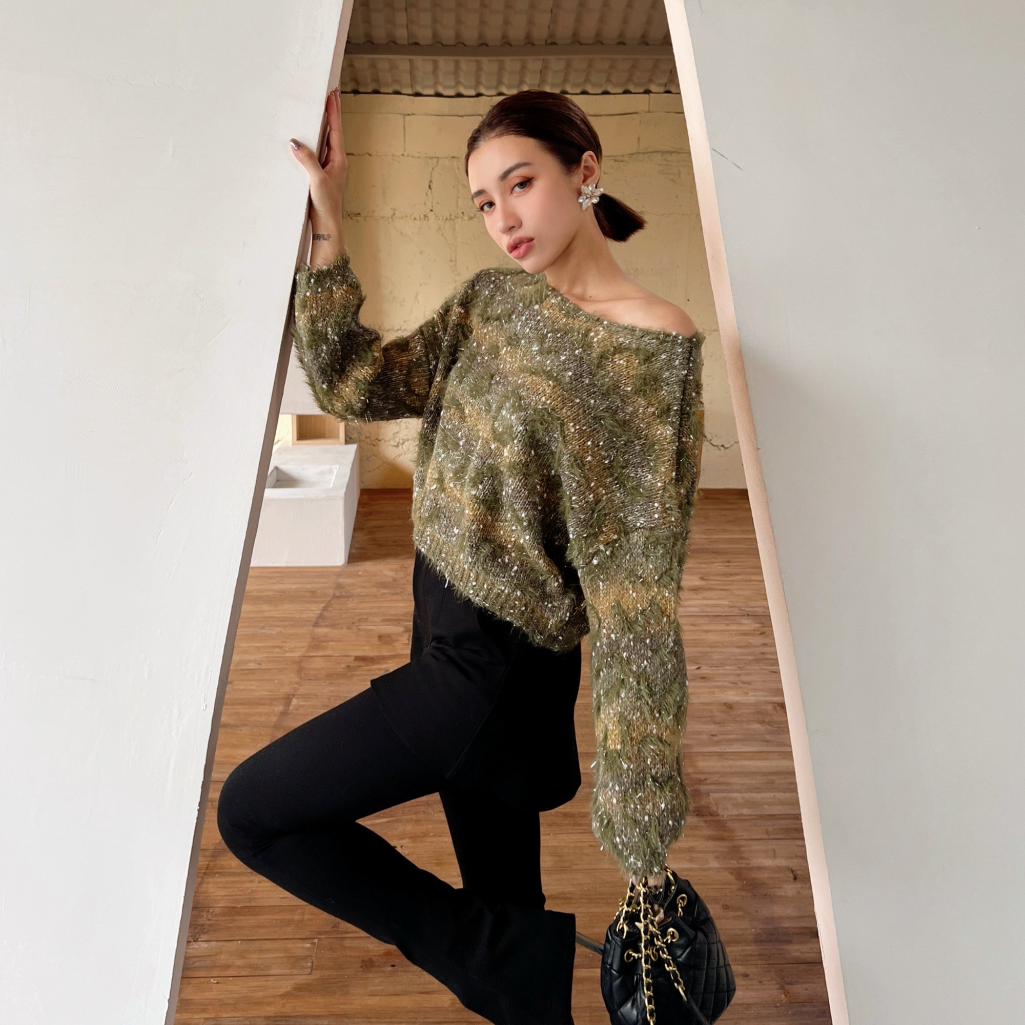 A woman wearing a green and gold patterned sweater and black pants is posing in a doorway, holding a black purse.