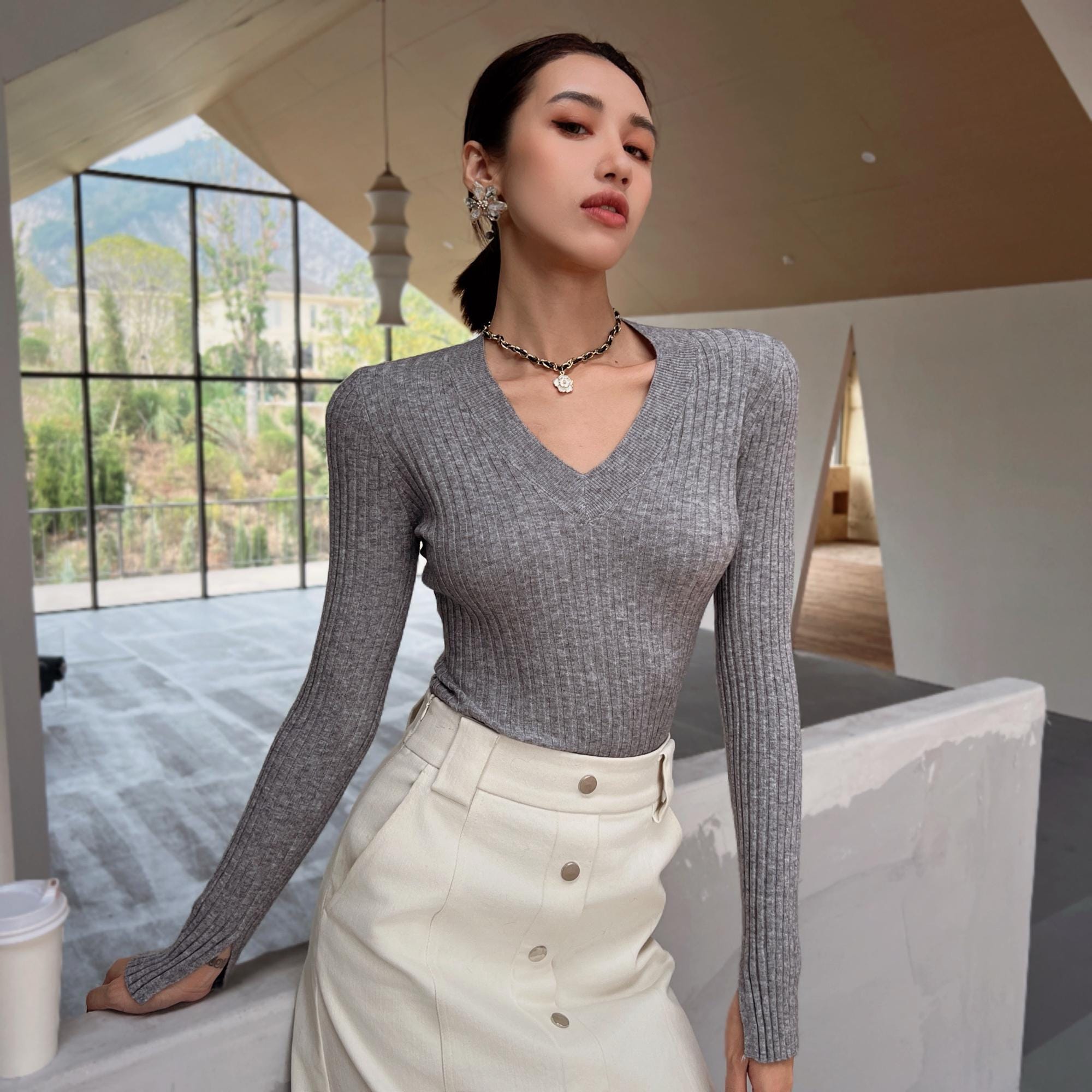 A woman wearing a gray sweater and a white skirt stands in a room with a large window overlooking a mountainous landscape.