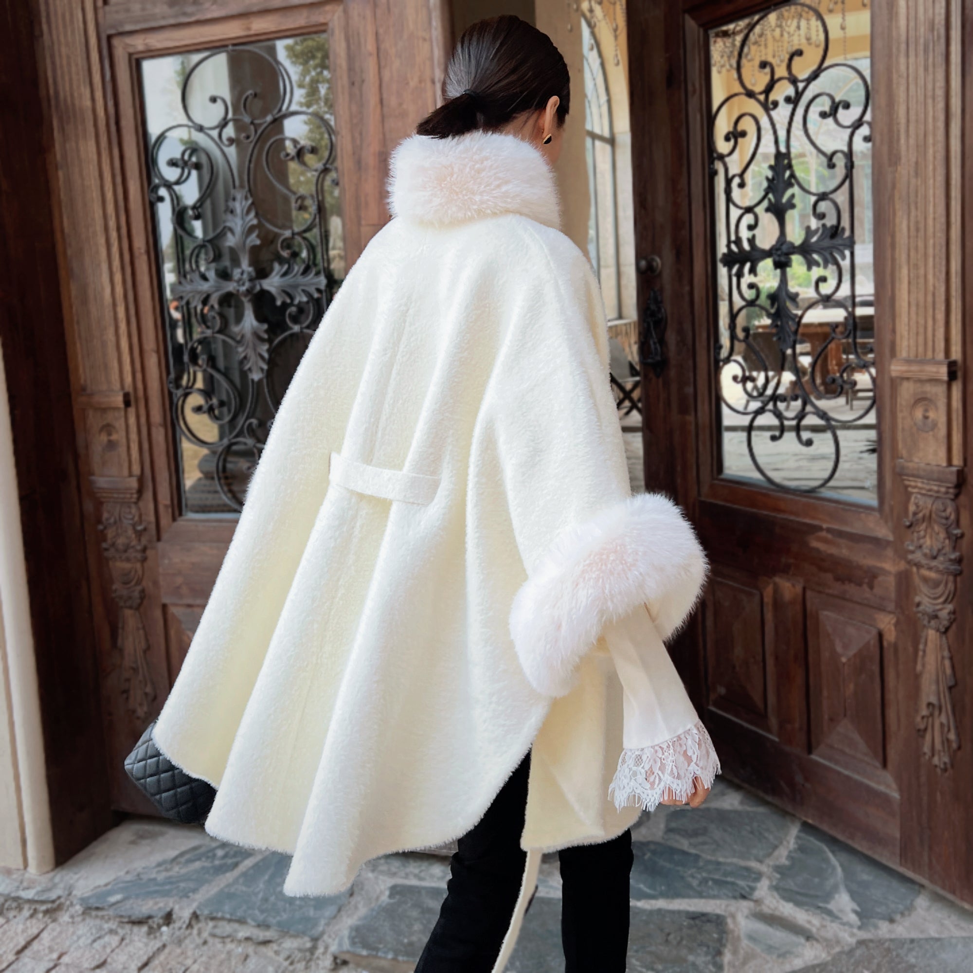 A woman wearing a white fur-trimmed cape walks past a wooden door with wrought iron accents.