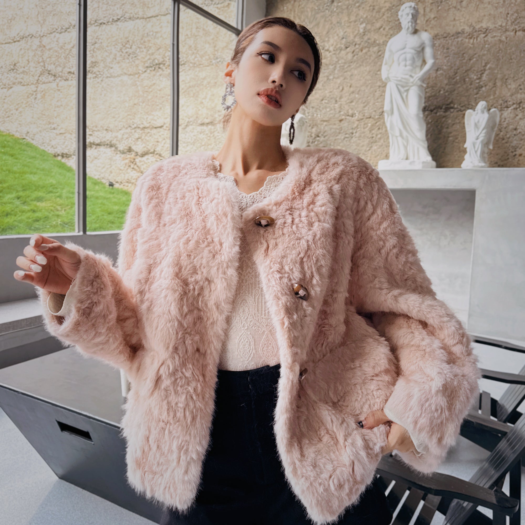 A woman wearing a pink fur coat stands in front of a window, with a statue visible in the background.