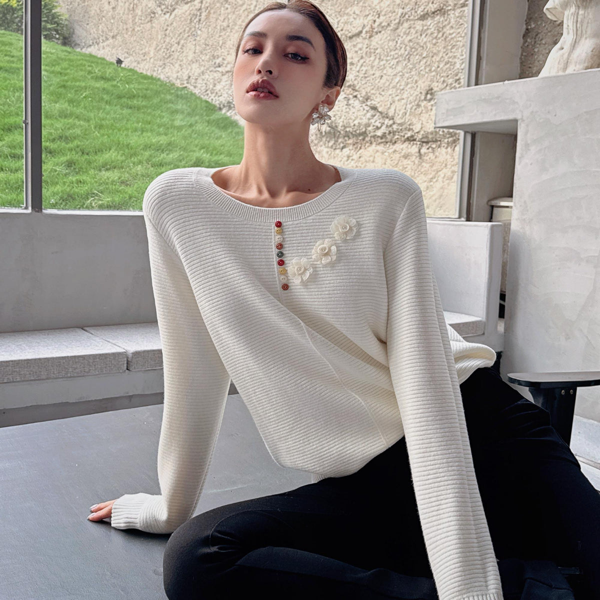 A woman wearing a white sweater with colorful flowers on the front is sitting on the floor, looking directly at the camera.