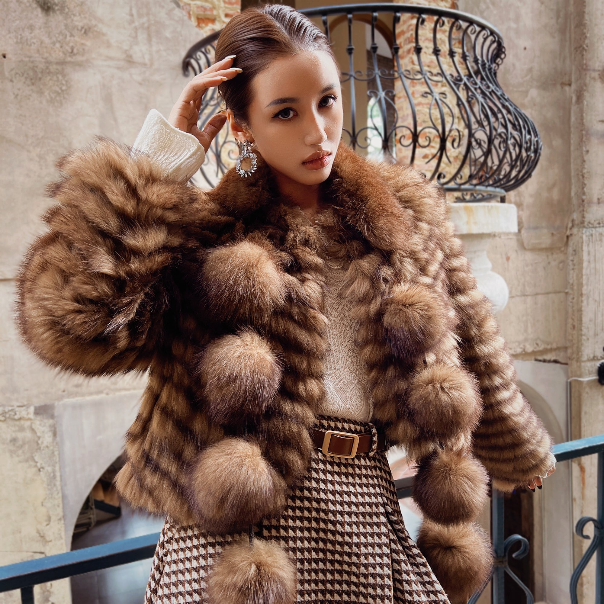 A woman wearing a fur coat and a patterned skirt stands in front of a wrought iron fence.