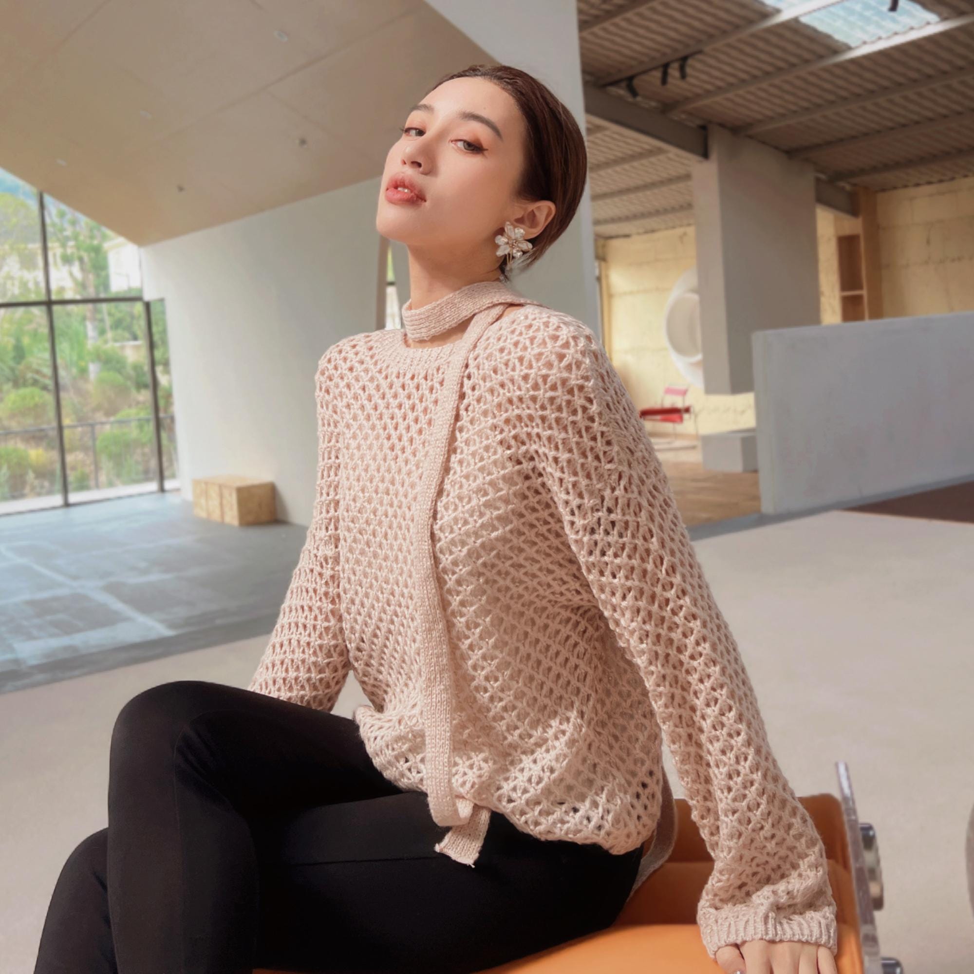 A woman wearing a beige sweater and black pants sits on a chair in an indoor setting, looking off to the side with a thoughtful expression.