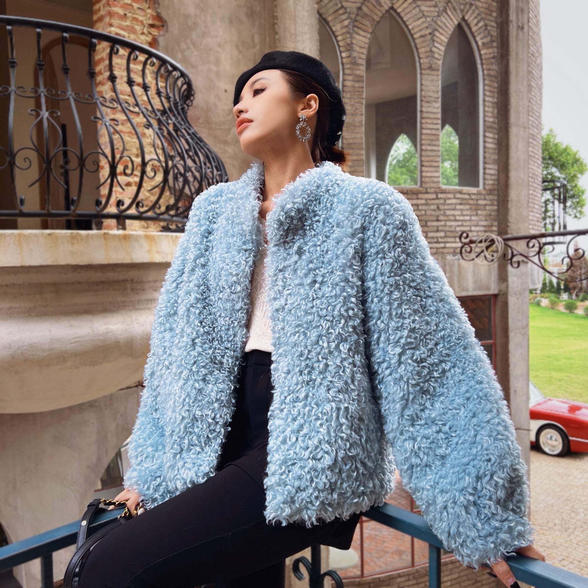 A woman wearing a blue fur coat and black pants is sitting on a balcony, looking off into the distance.