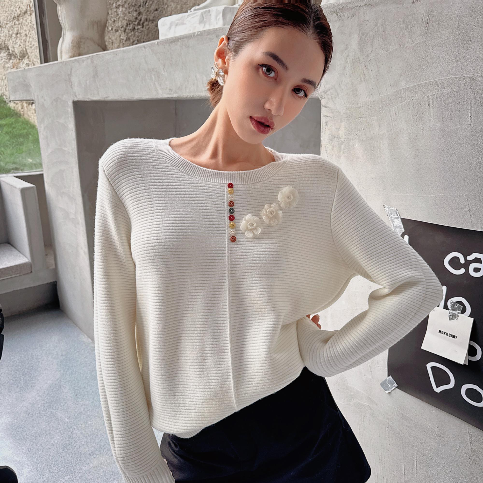 A woman wearing a white sweater with a floral design and a red and orange flower on the front, posing in an indoor setting.